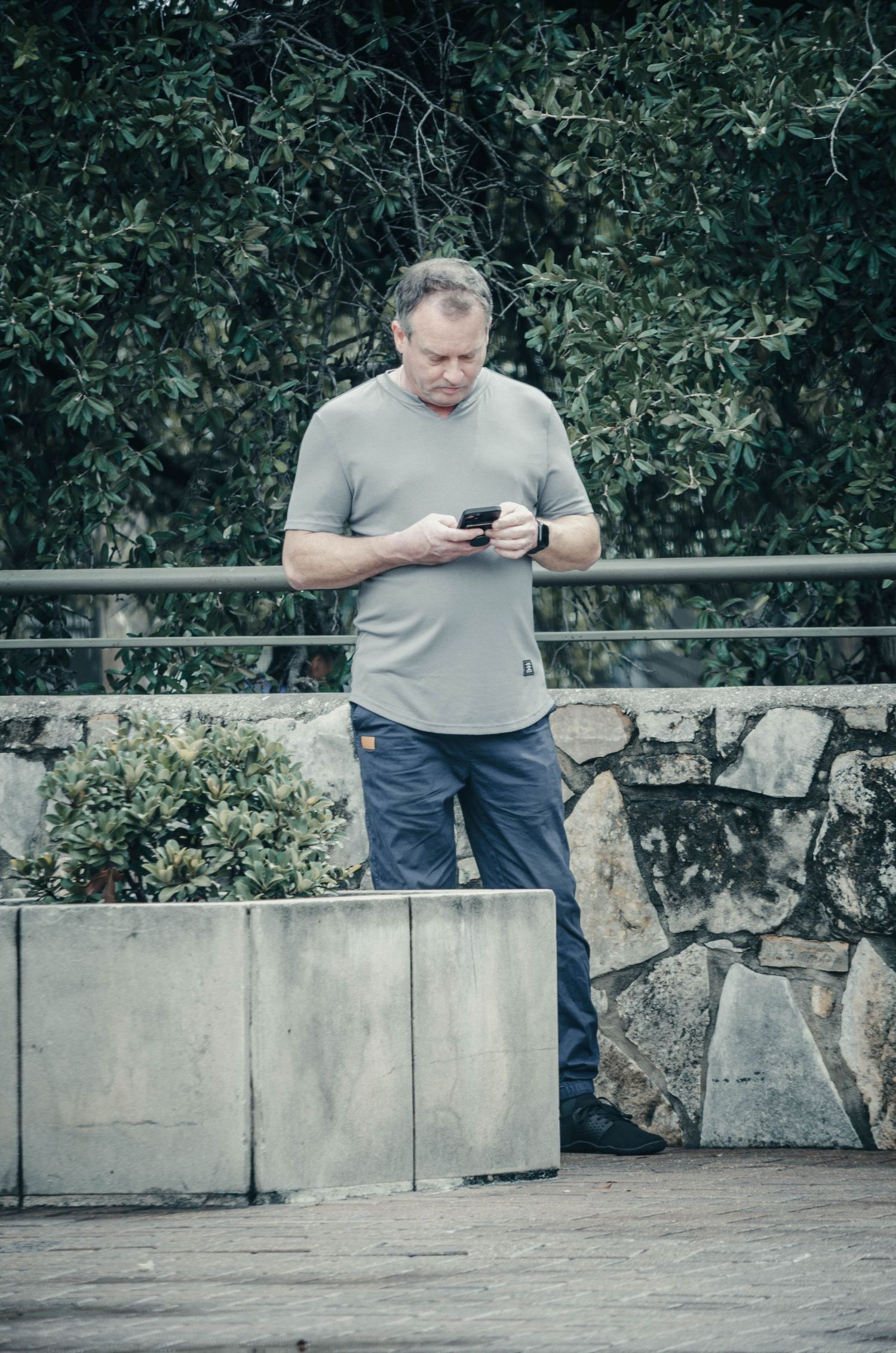 Middle-aged man in gray t-shirt and blue pants checks his smartphone outdoors near a stone wall.