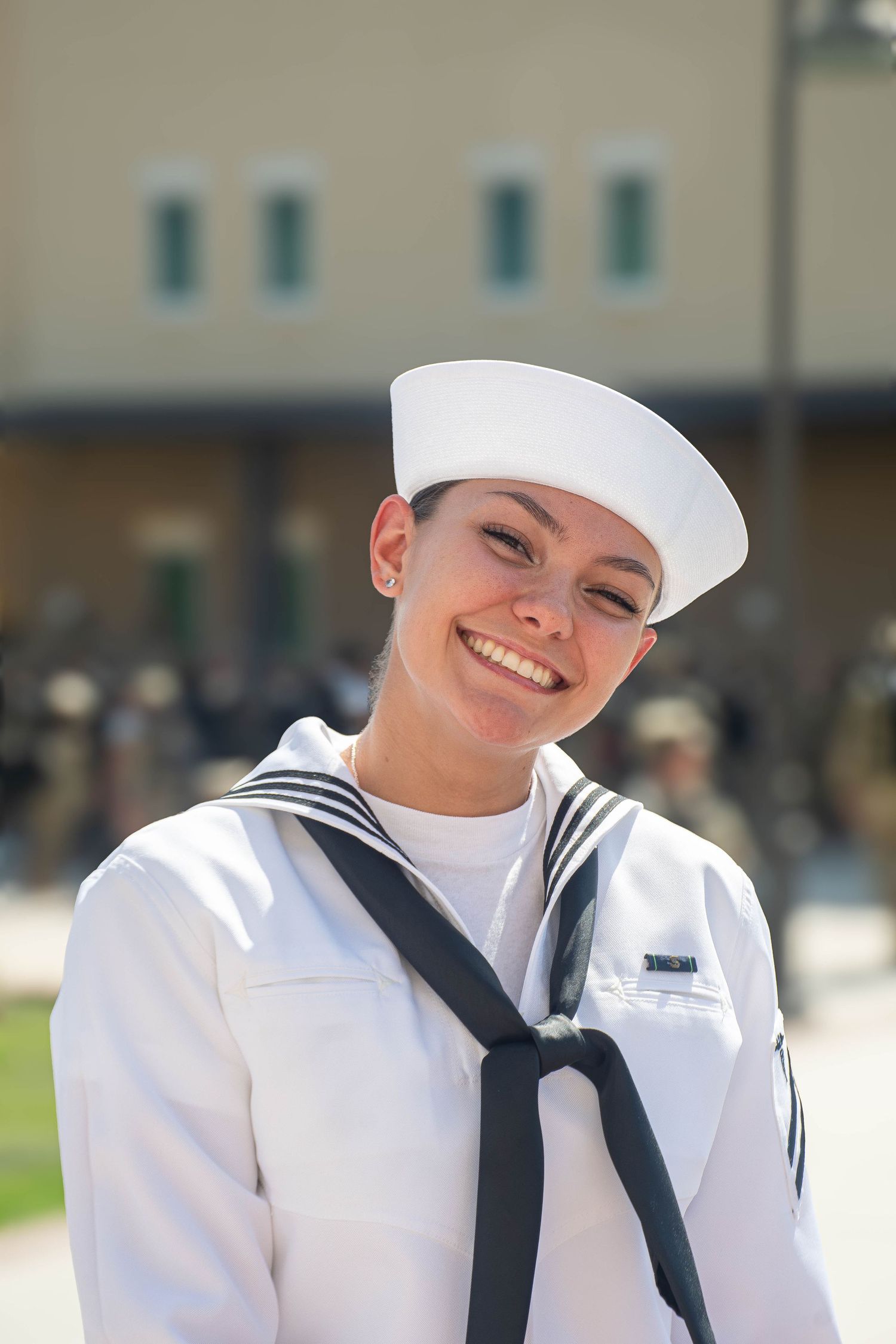 Young female Navy sailor in dress whites smiles brightly at outdoor military graduation ceremony.