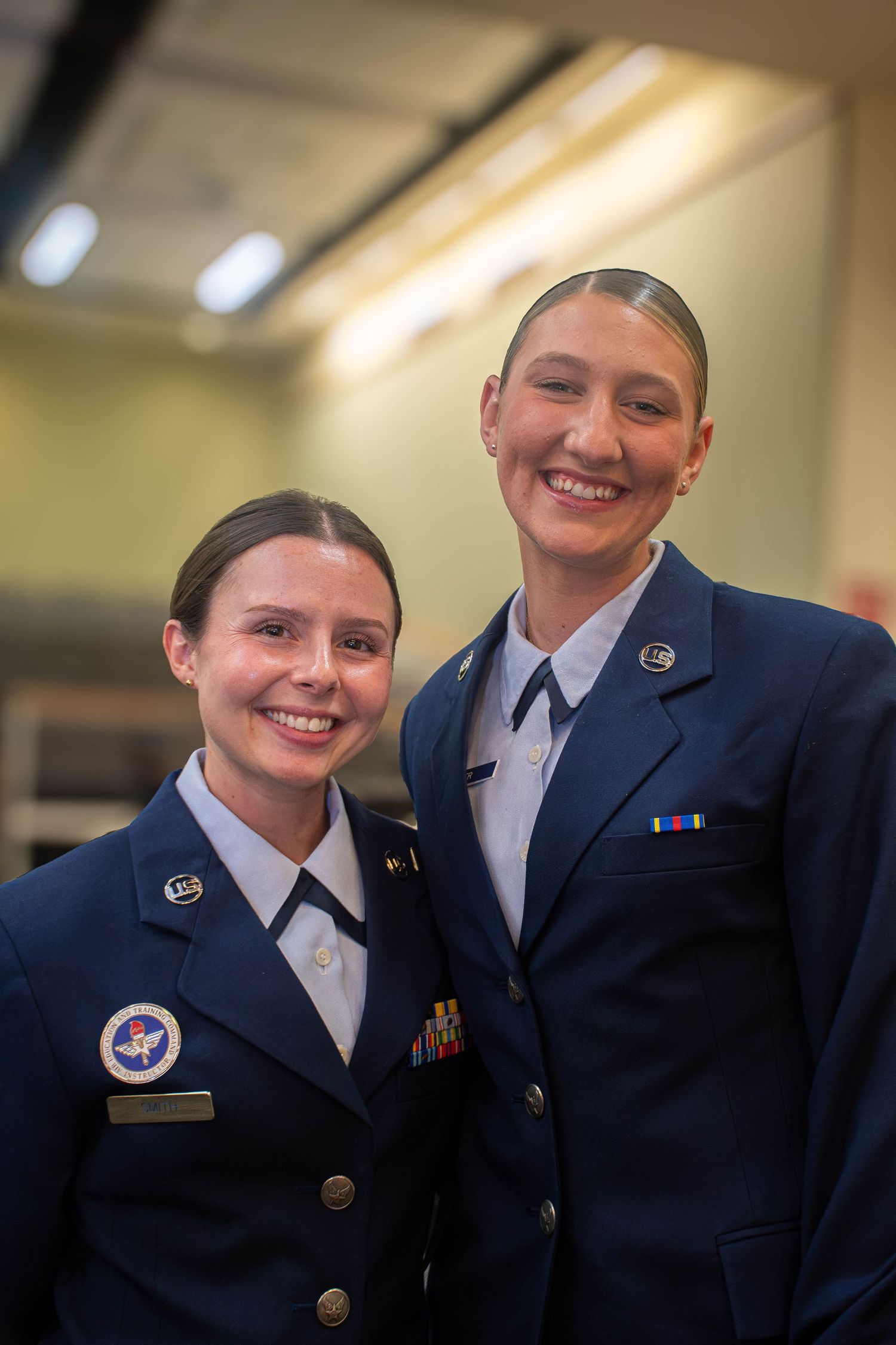Two smiling female Air Force cadets in blue dress uniforms pose together indoors.