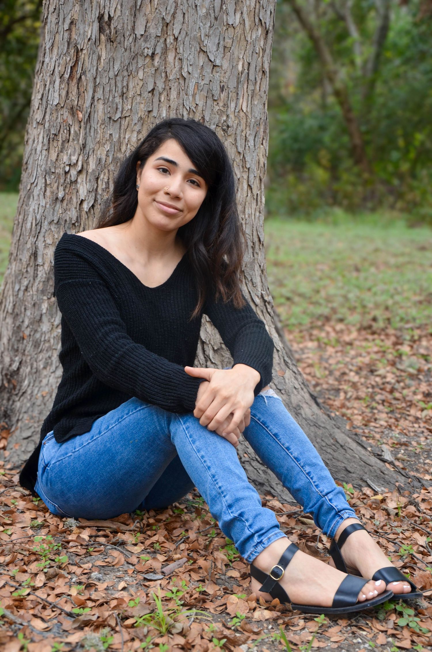 Young woman in black sweater and jeans sitting against tree trunk on autumn leaves in park.
