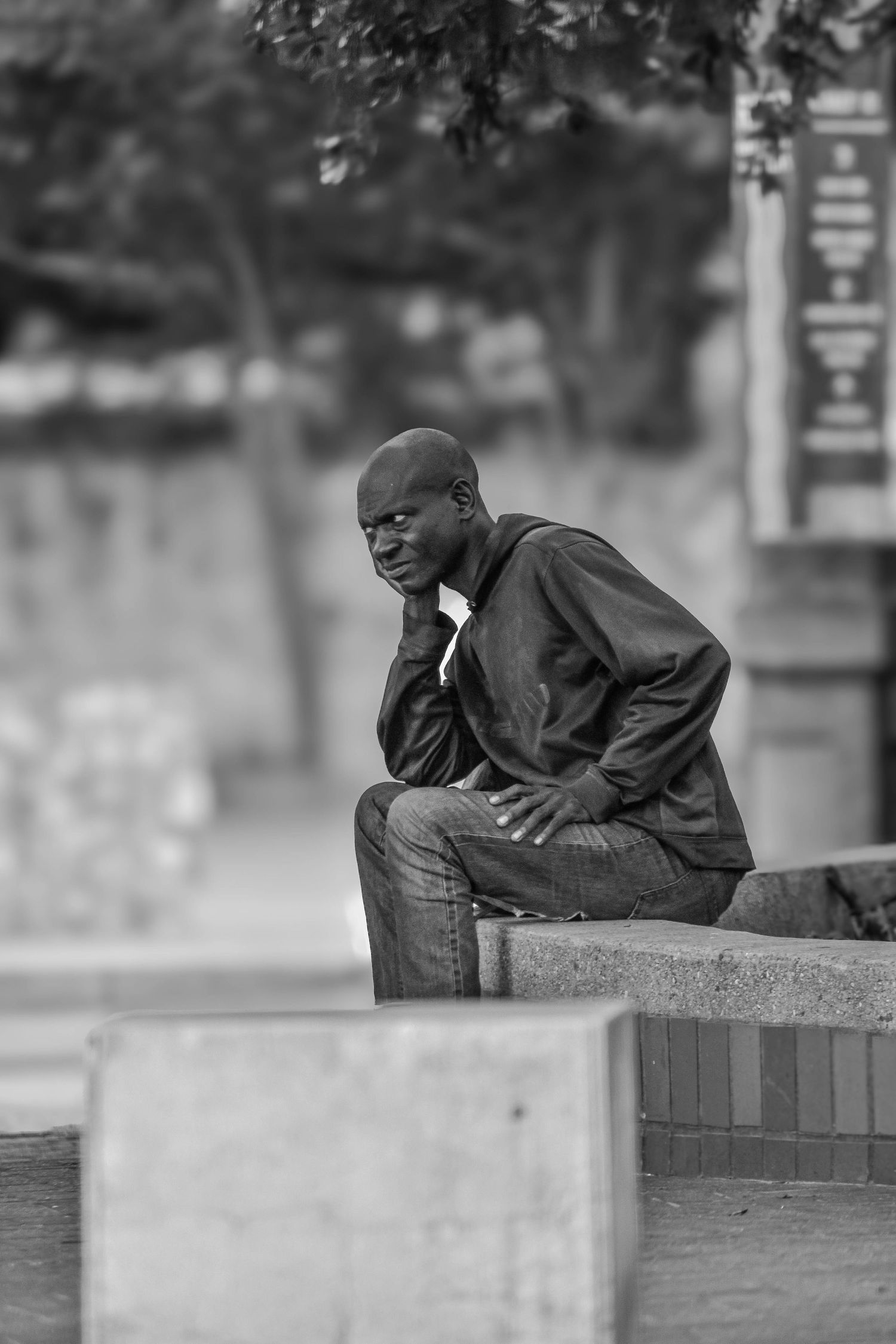Black and white photo of a young man sitting on a concrete ledge outdoors, resting his chin on his hand in a thoughtful pose.