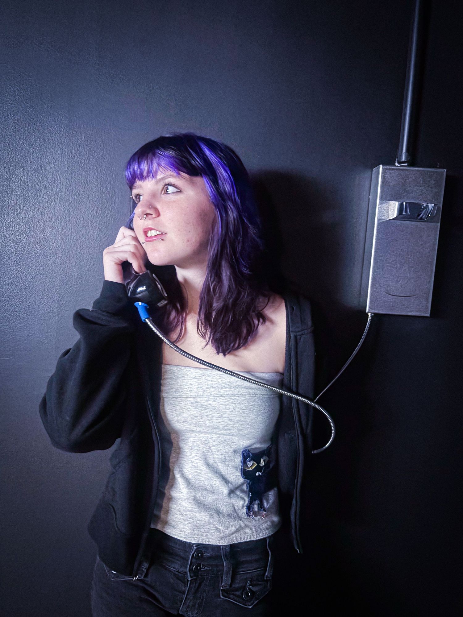 Young woman with purple hair talking on a corded payphone against a dark wall wearing a black hoodie.