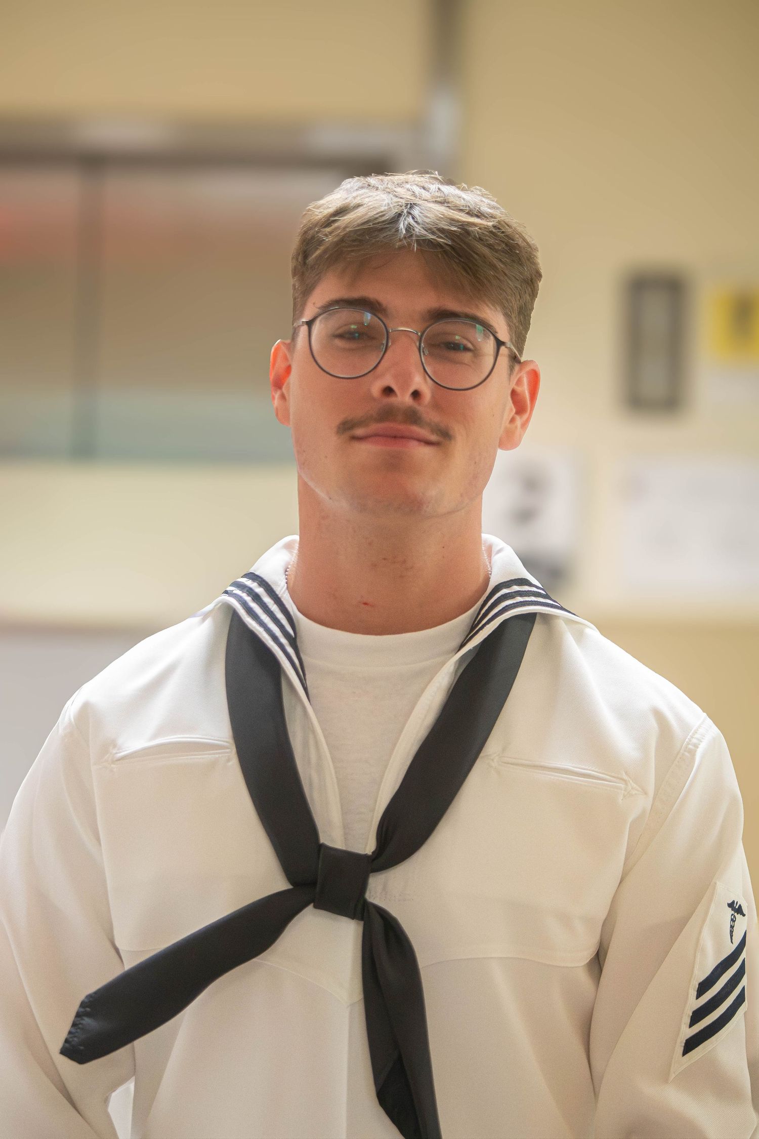 Young male Navy sailor with glasses and mustache in dress white uniform poses indoors.