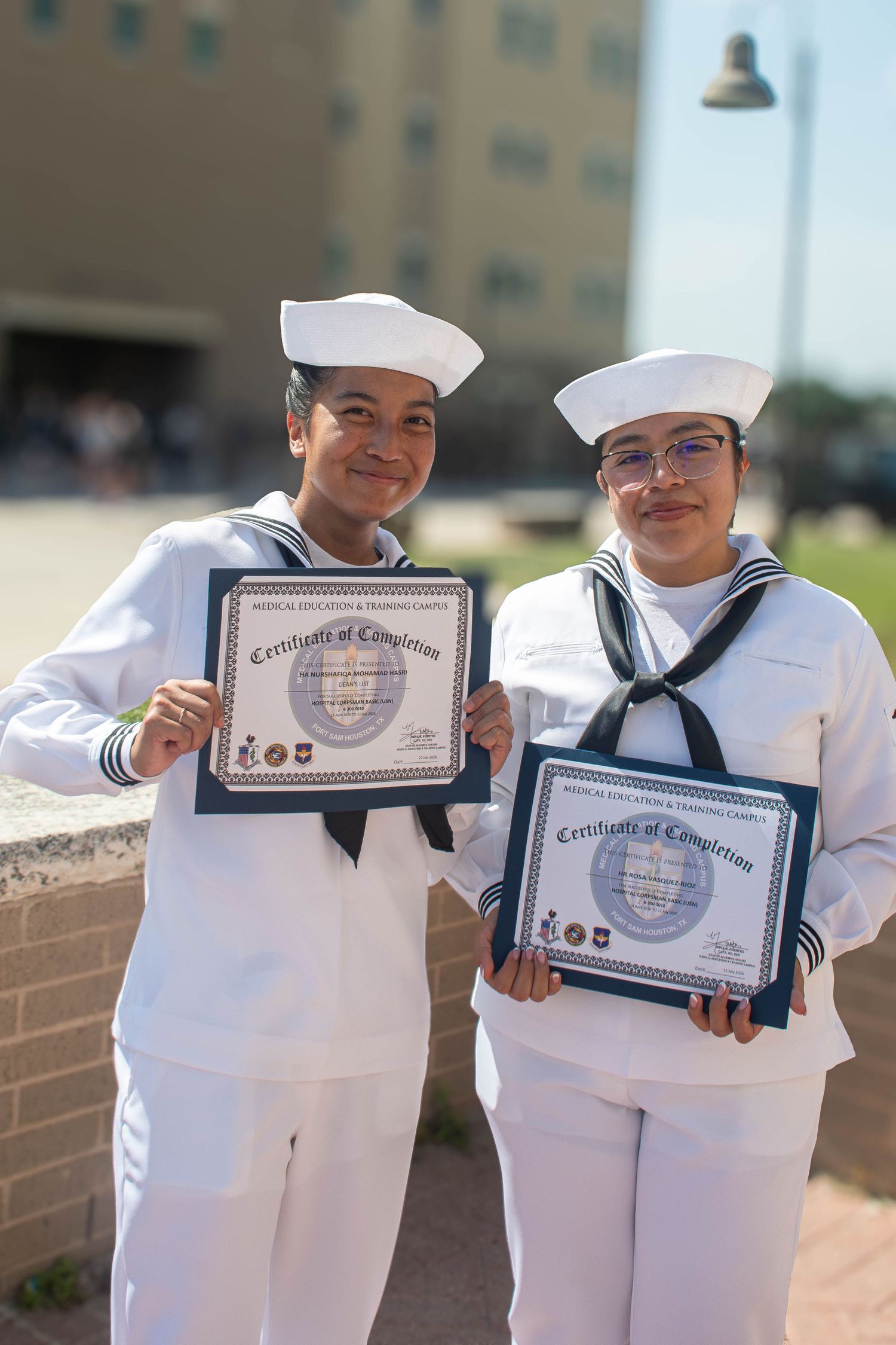 Two female Navy sailors proudly display Certificates of Completion in dress white uniforms outdoors.