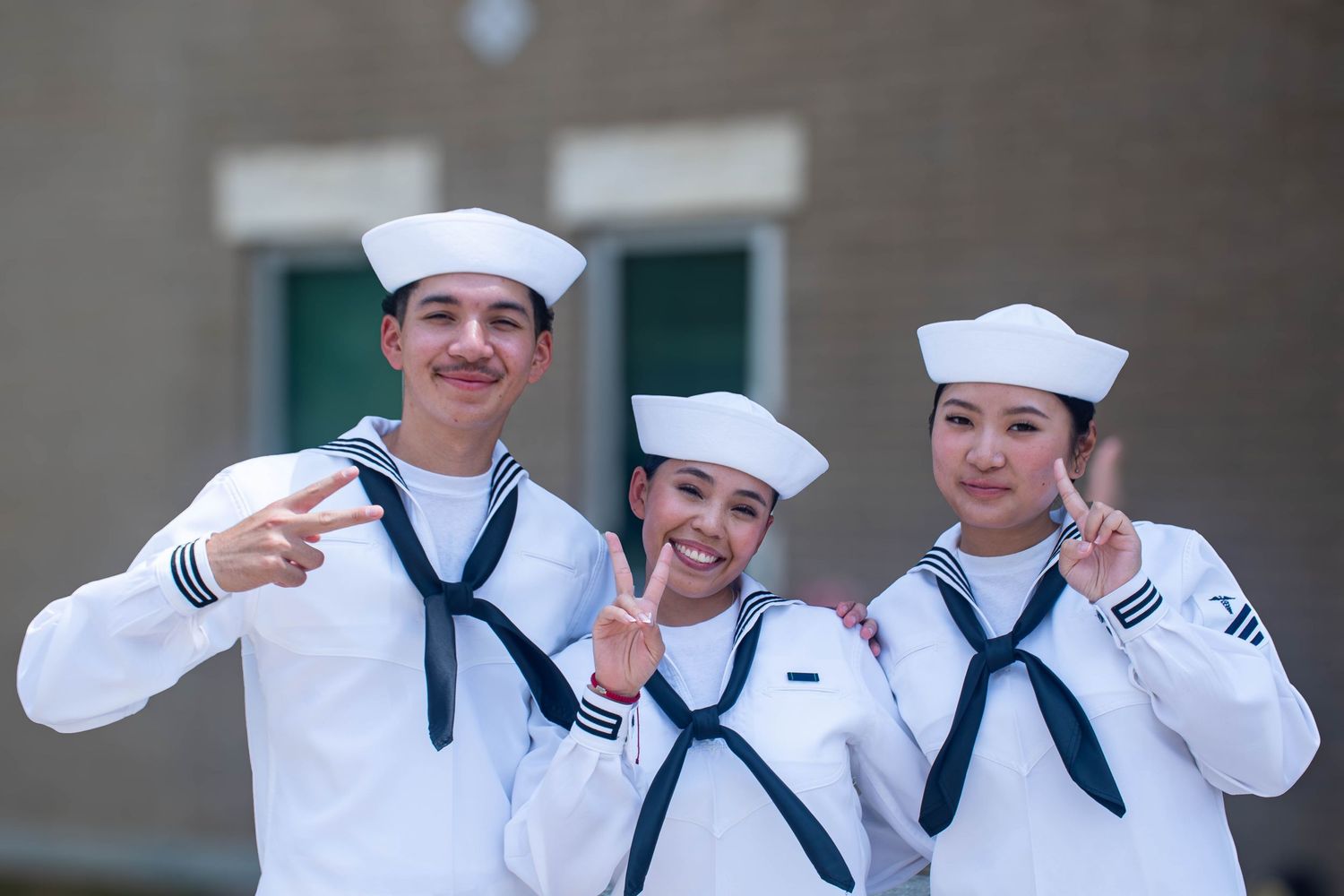 Three smiling Navy sailors in white uniforms make peace signs outdoors.