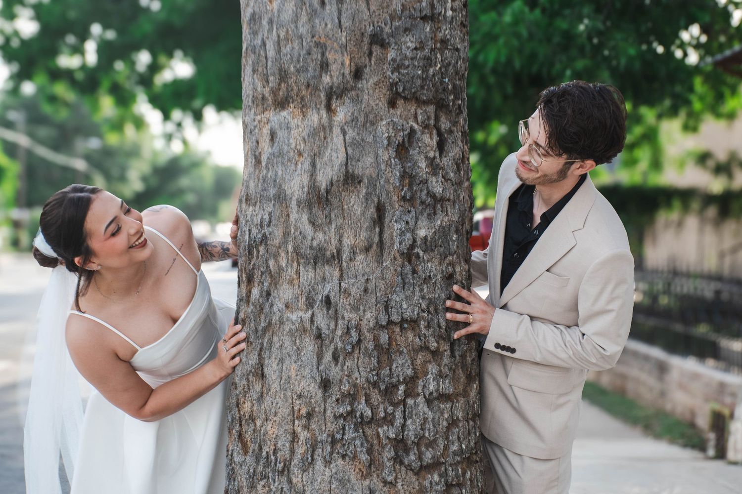 Playful bride and groom peek around opposite sides of a large tree trunk outdoors on their wedding day.