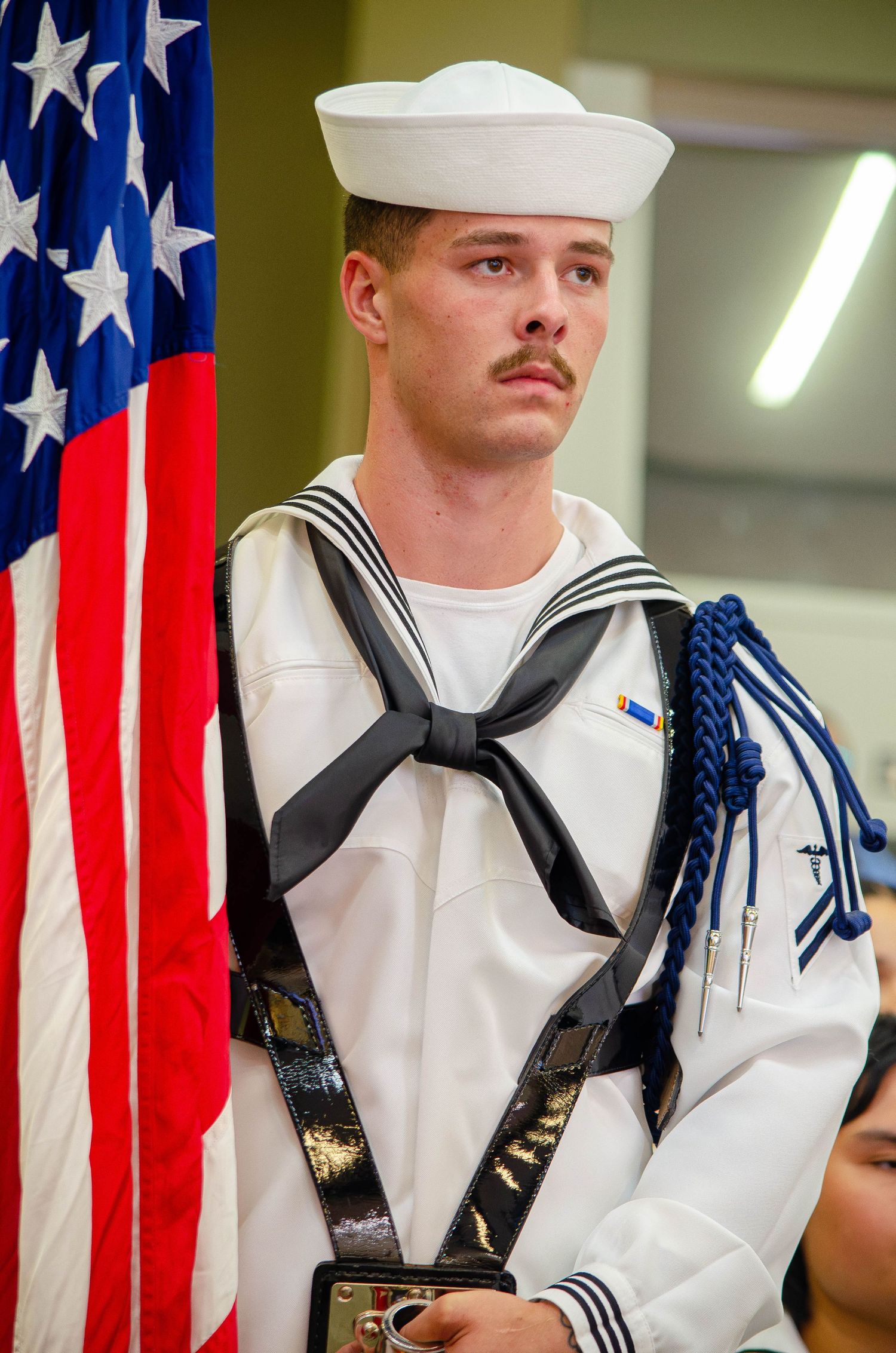Male Navy sailor in dress whites holding US flag at formal military ceremony.