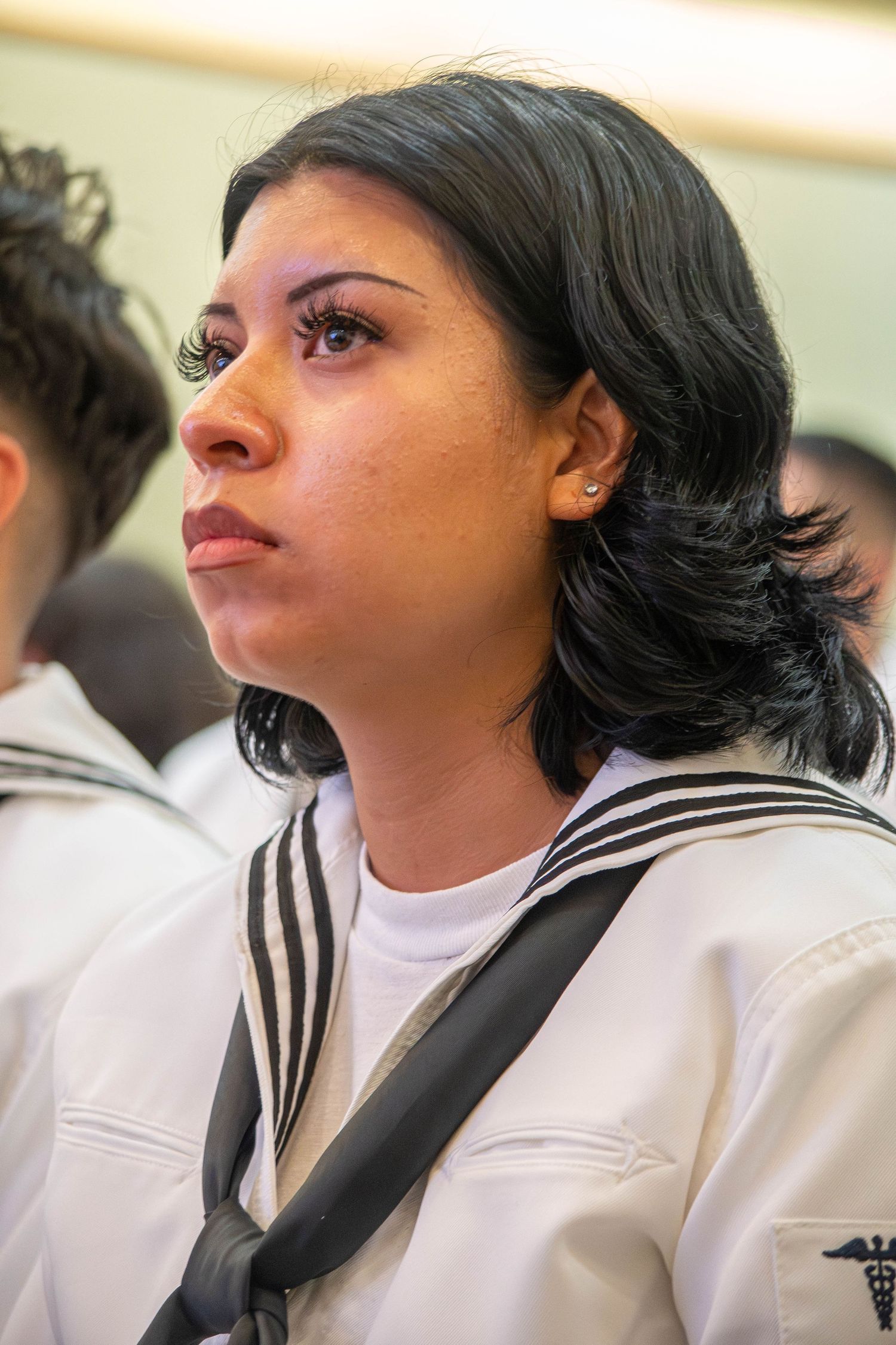 Female Navy sailor with black hair in white uniform with caduceus insignia gazing upward.