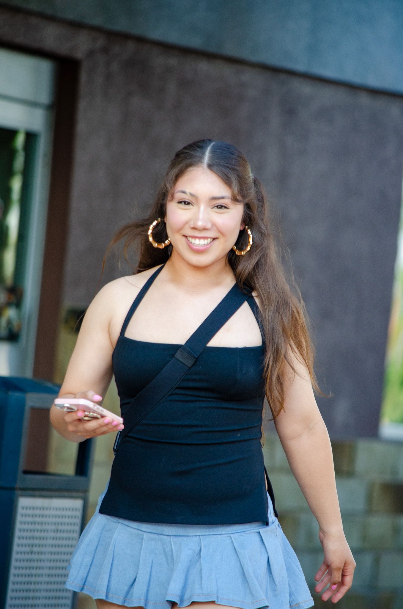 Young woman in black halter top and blue skirt smiling while holding a phone outdoors.