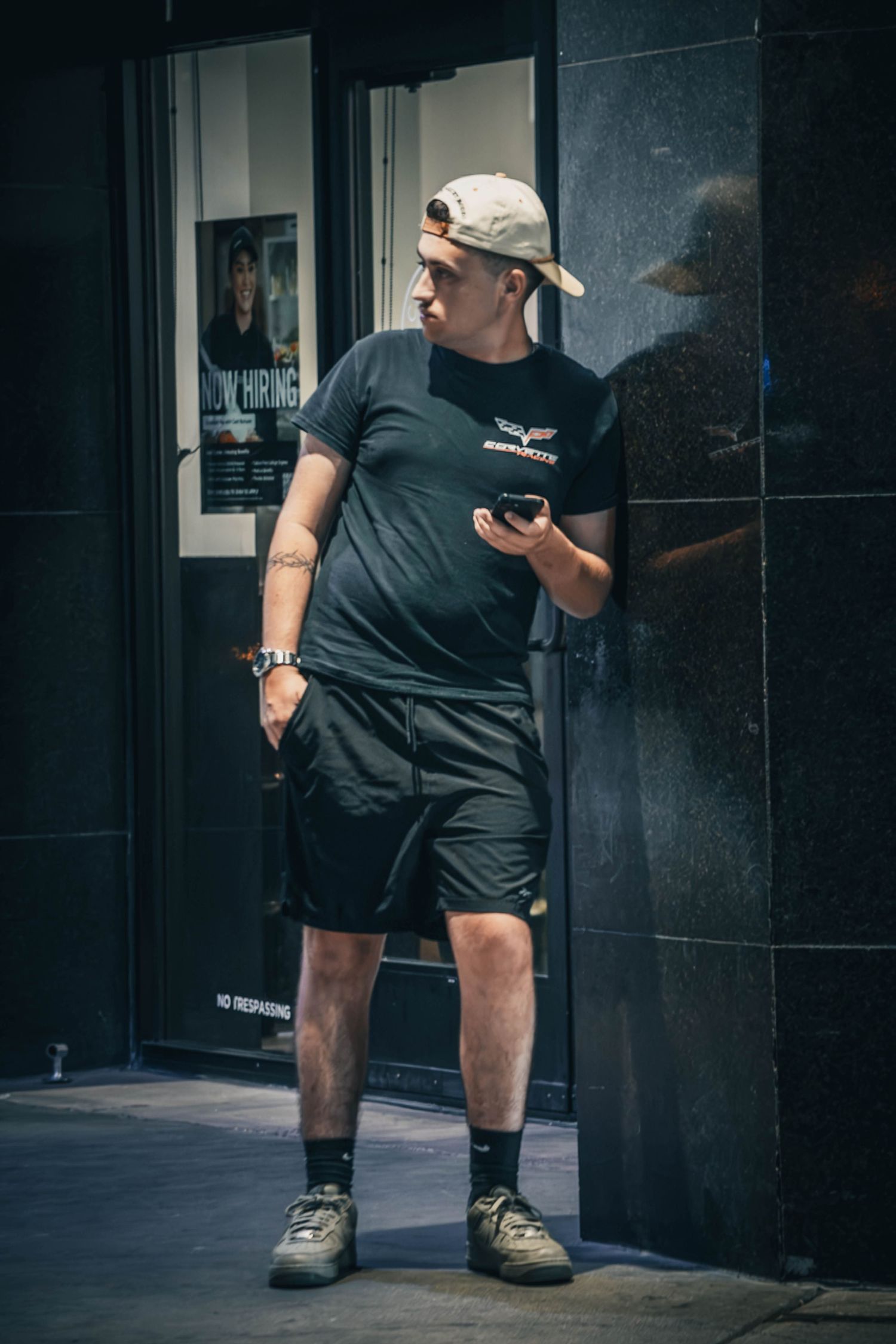 Man in black t-shirt, shorts, and cap leans against a dark wall while looking at his phone outdoors.