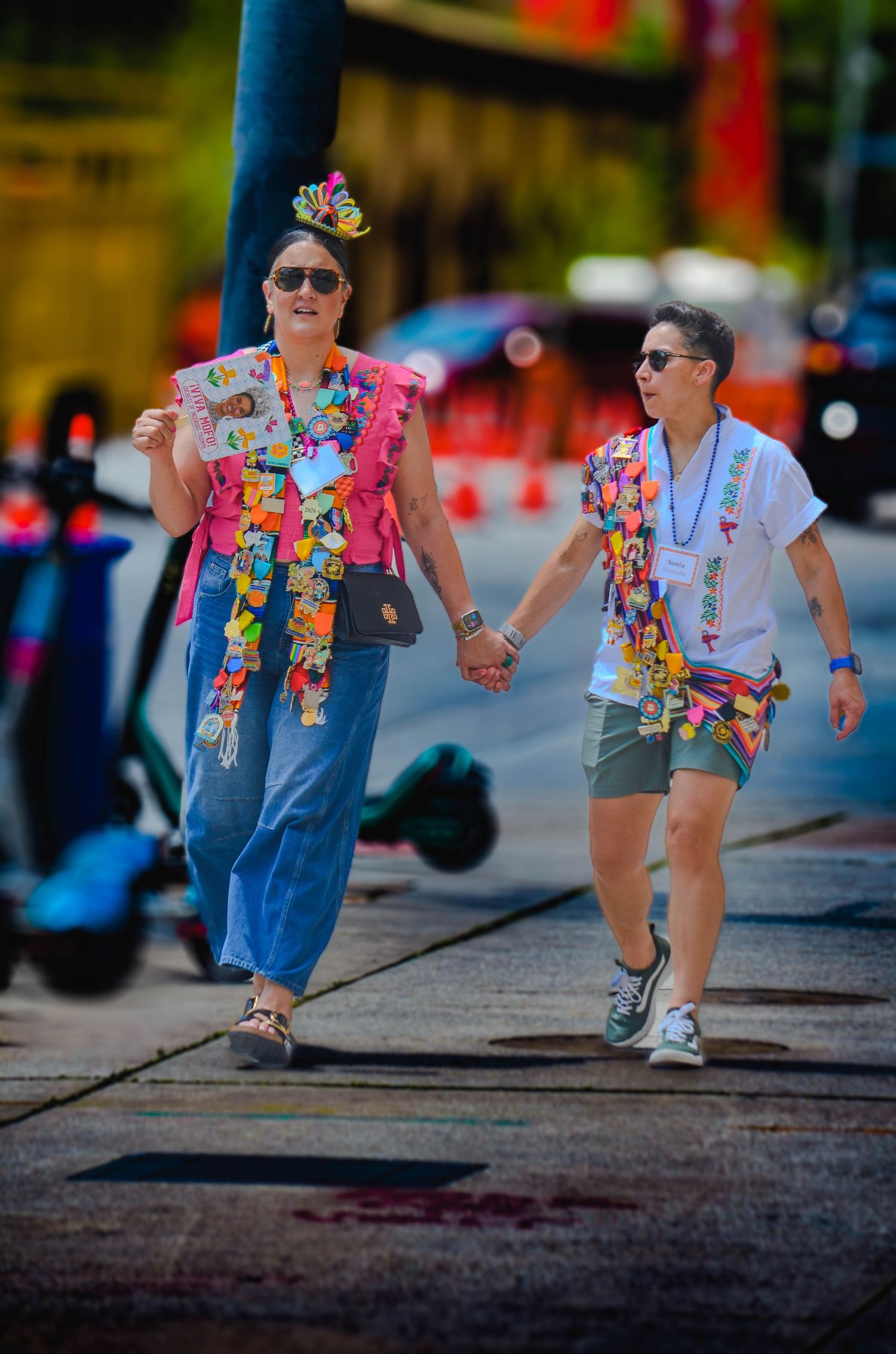 Two women in colorful festival attire holding hands while walking on city sidewalk.
