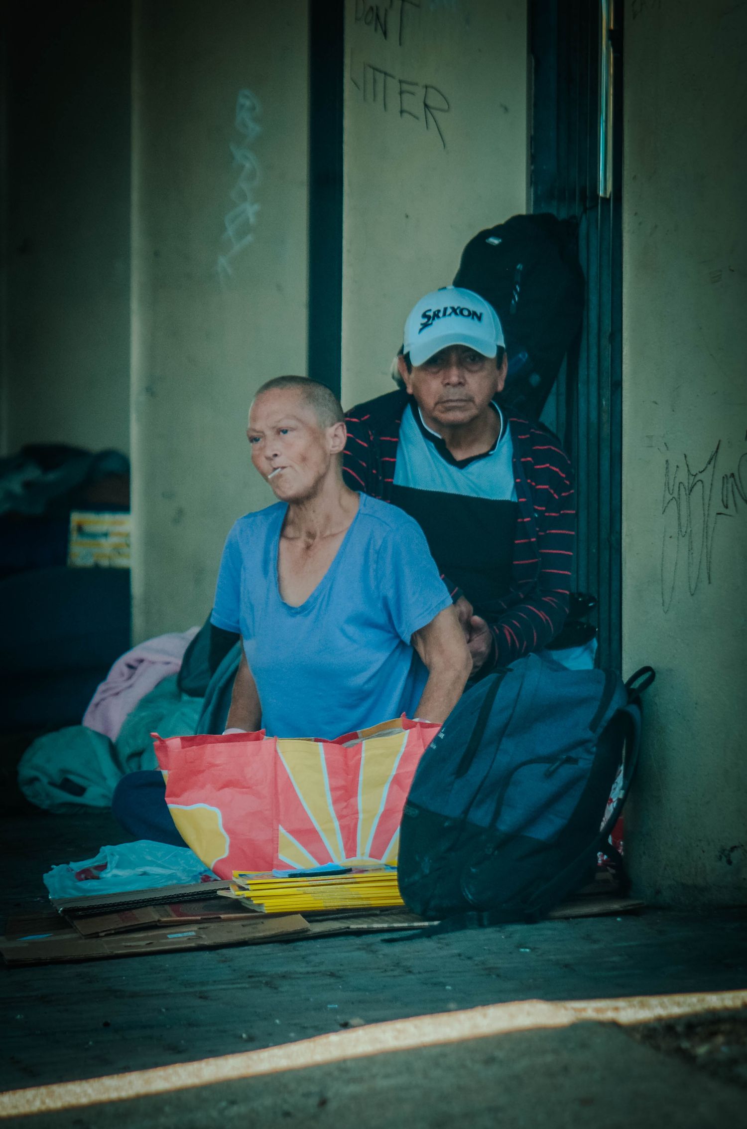 Two people sitting in a doorway with bags and colorful items in an urban street setting.