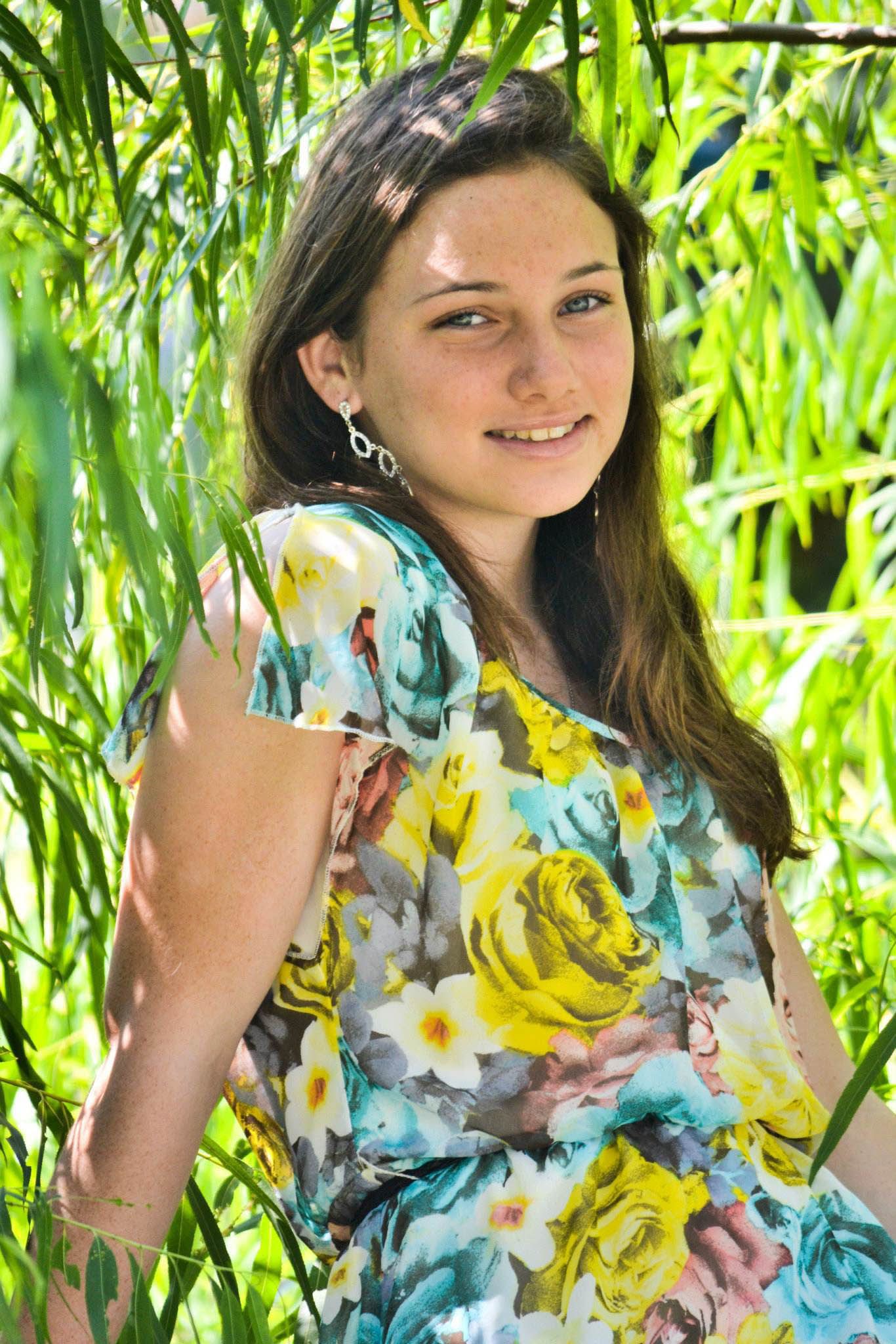 Smiling teenage girl in floral dress standing among green willow branches.