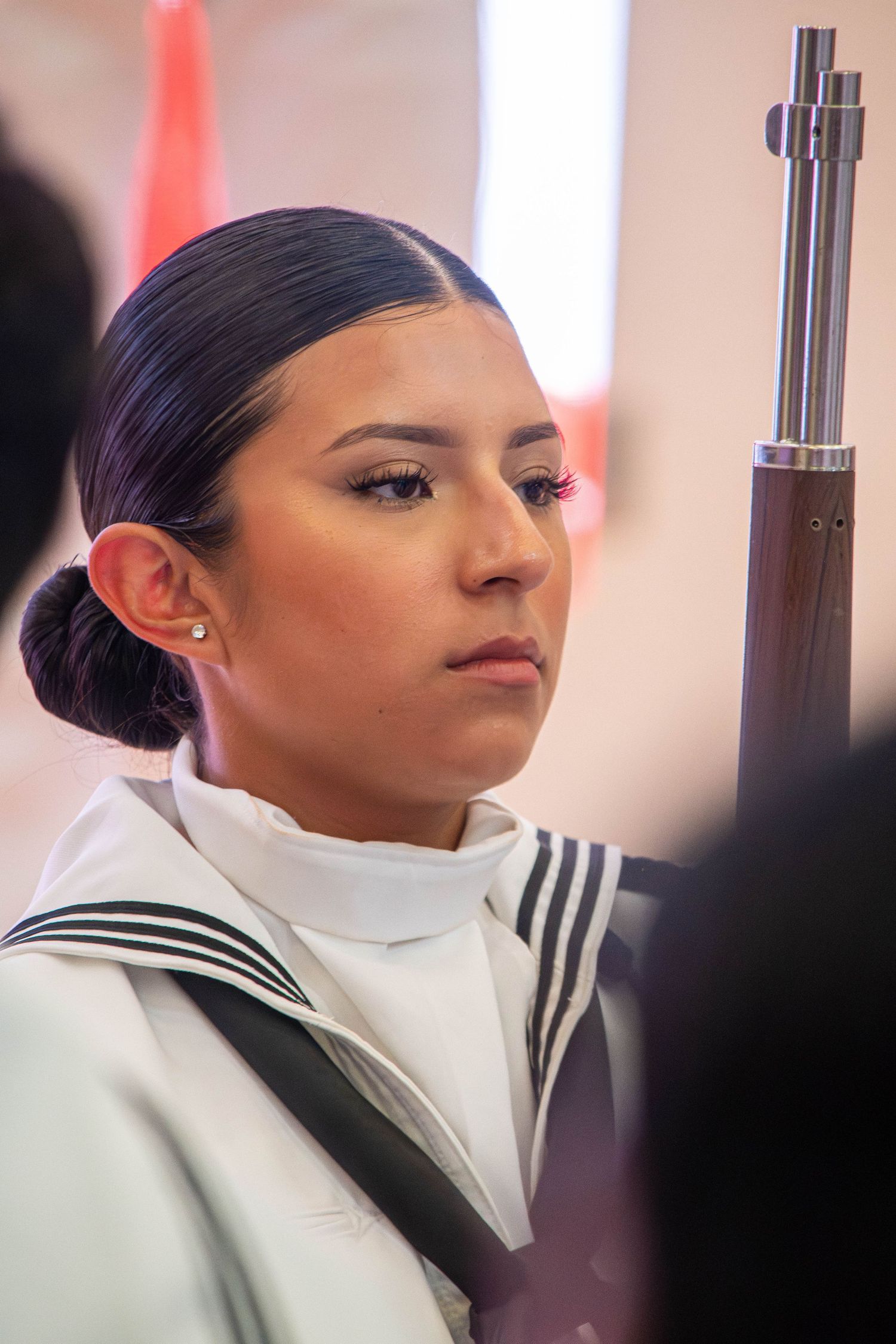 US Navy sailor in white dress uniform holding rifle during a formal color guard ceremony.