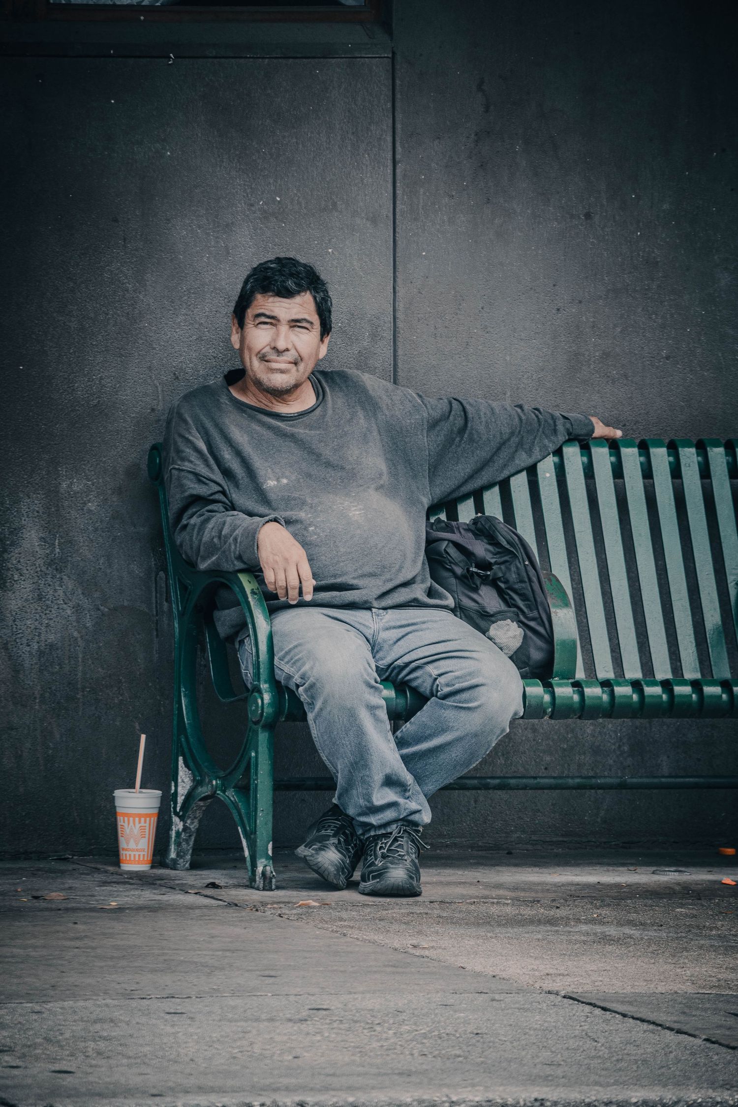 Smiling middle-aged man sitting relaxed on a green bench outdoors with a drink nearby.