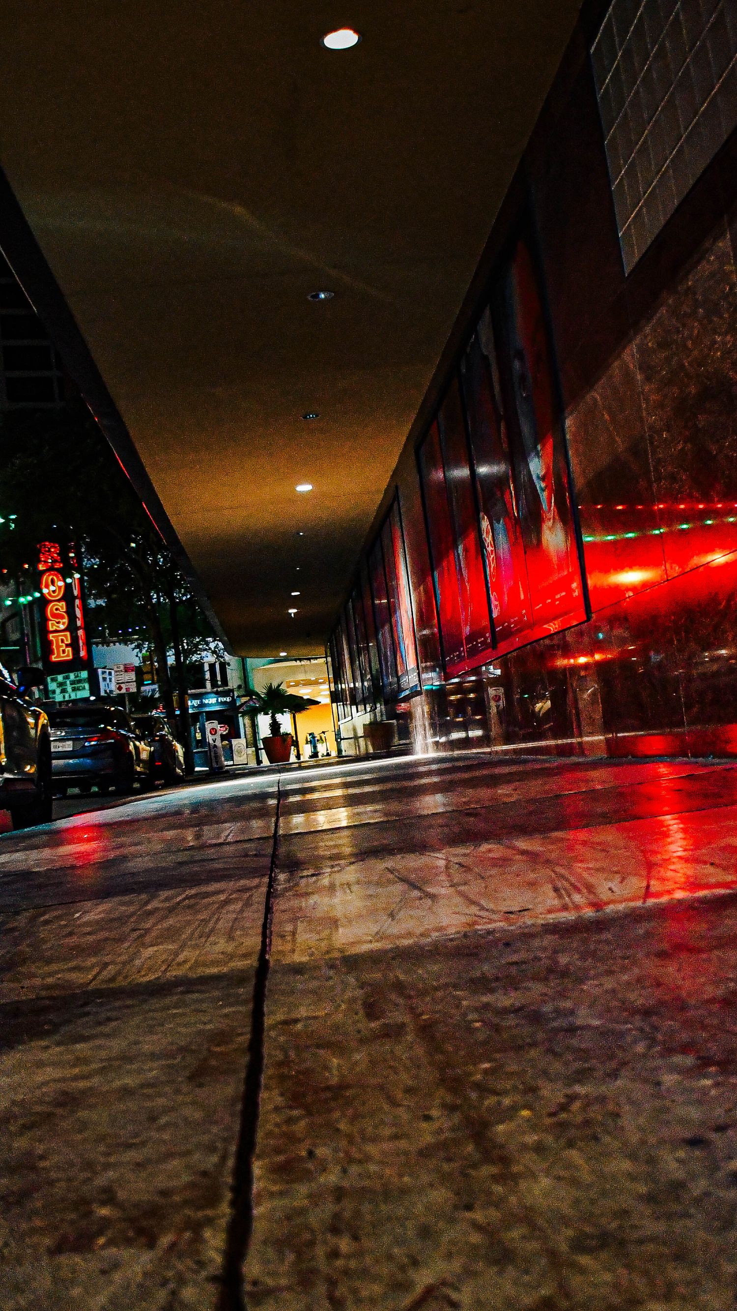 Low angle night shot of a wet urban sidewalk with colorful neon reflections on buildings.
