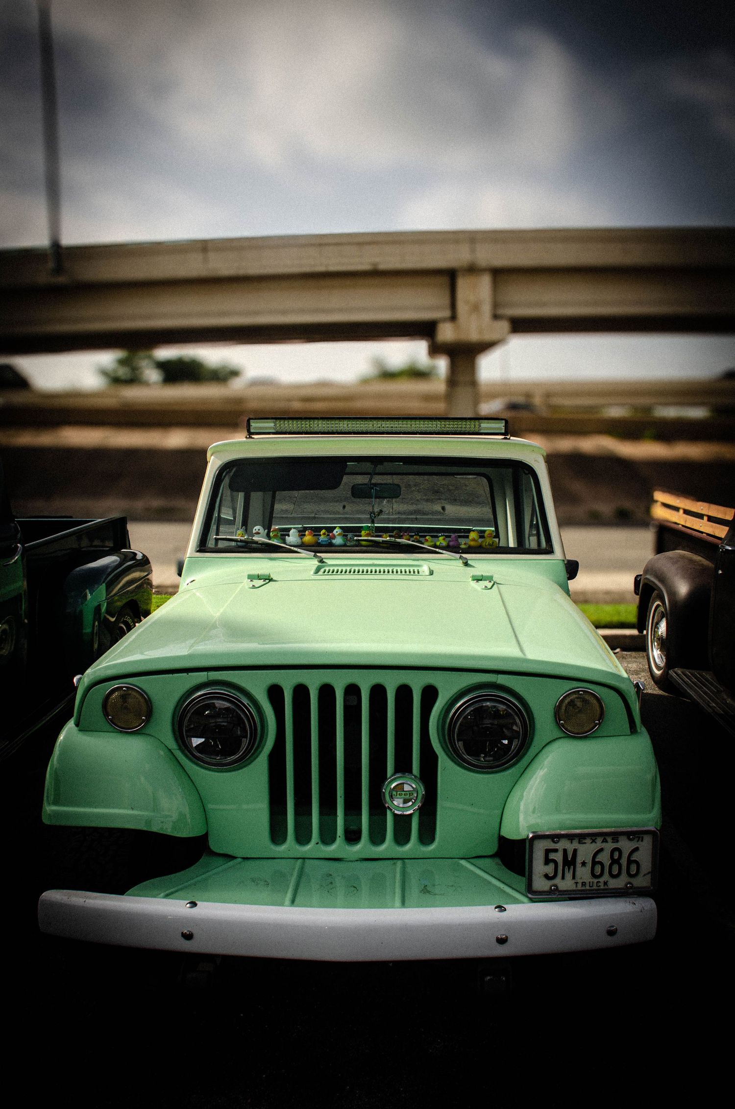 Vintage mint green Jeep Willys with license plate 5M-686 parked under overpass on cloudy day.