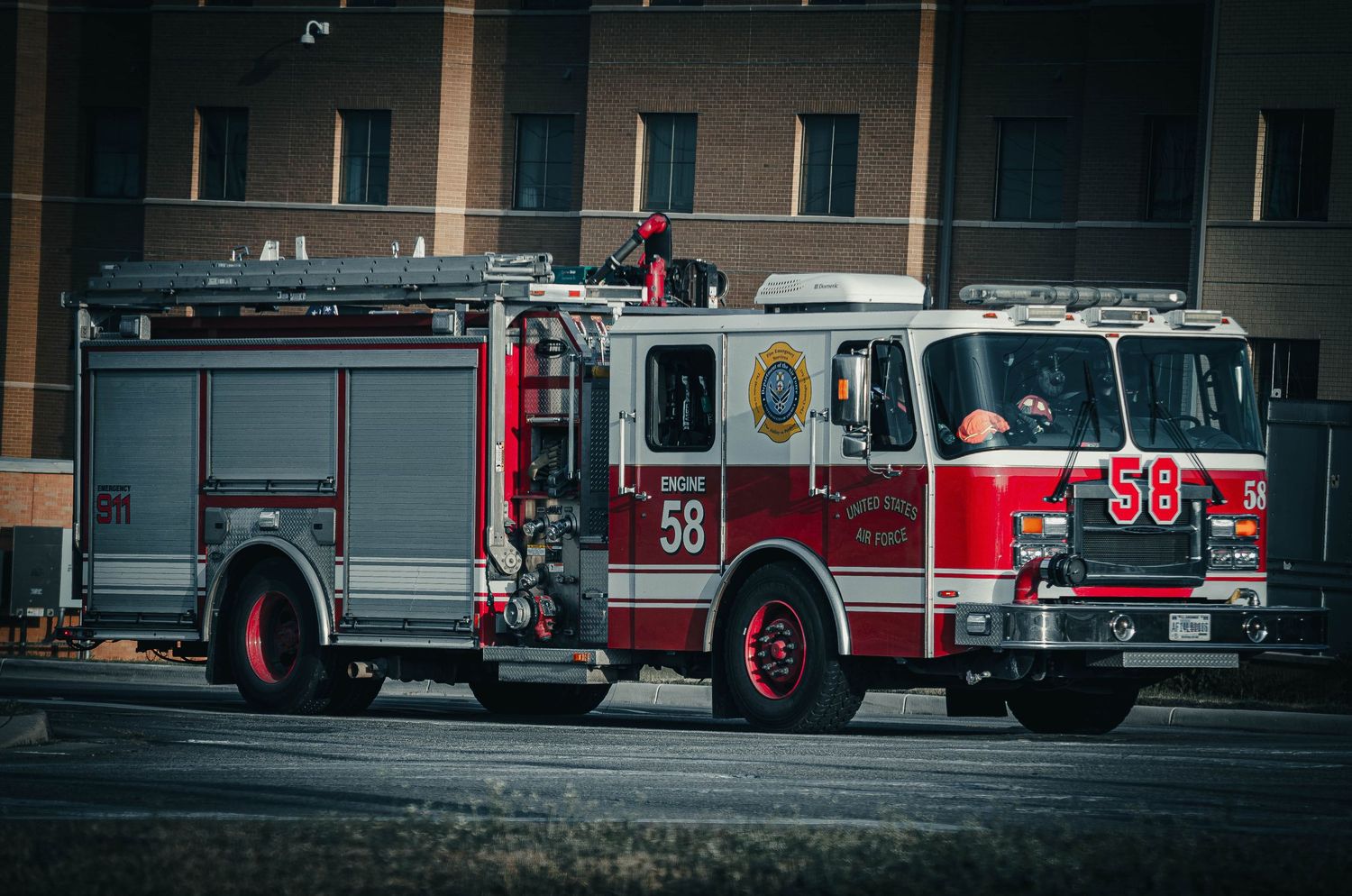 Red fire truck number 58 driving on a city street with a firefighter visible in the cab near a building.