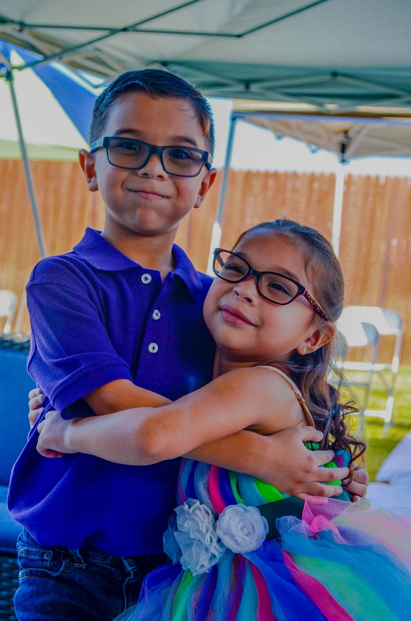 Two young siblings in glasses hugging at an outdoor party under a tent.