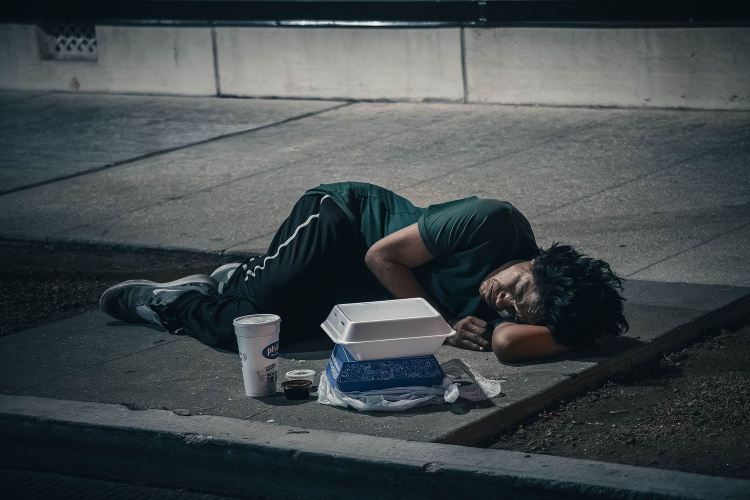 Person sleeping on a sidewalk curb at night with food containers and a cup nearby, depicting urban homelessness.