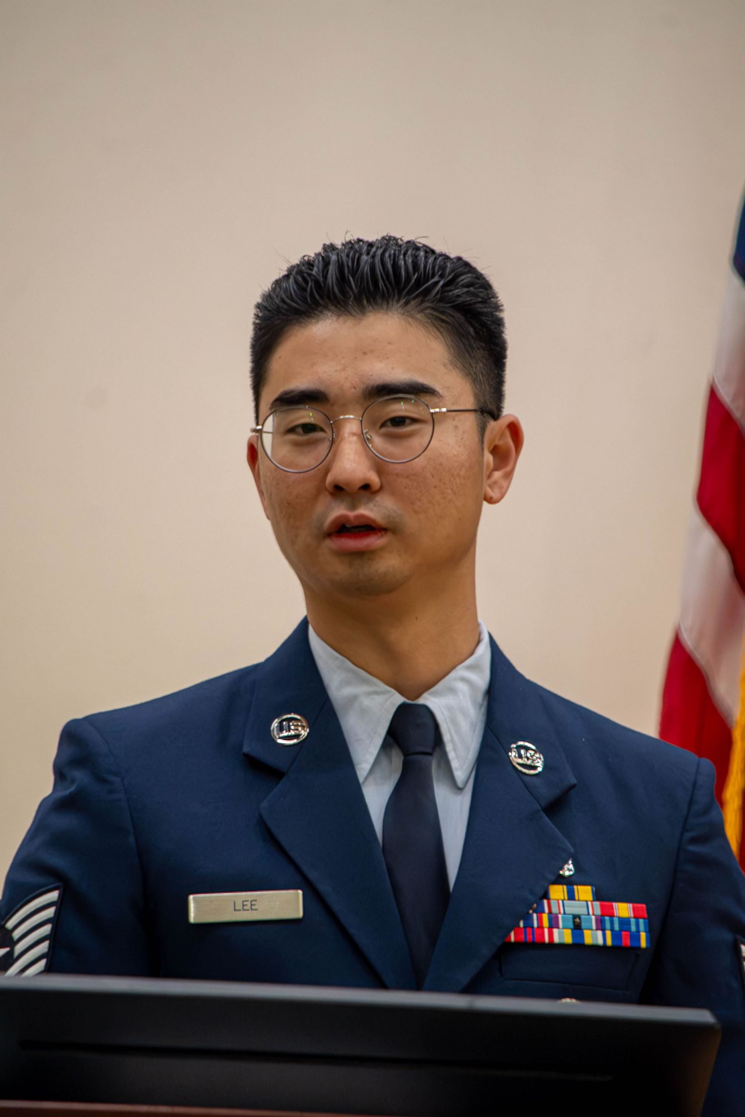 Male Air Force servicemember in blue dress uniform with ribbons stands at podium.