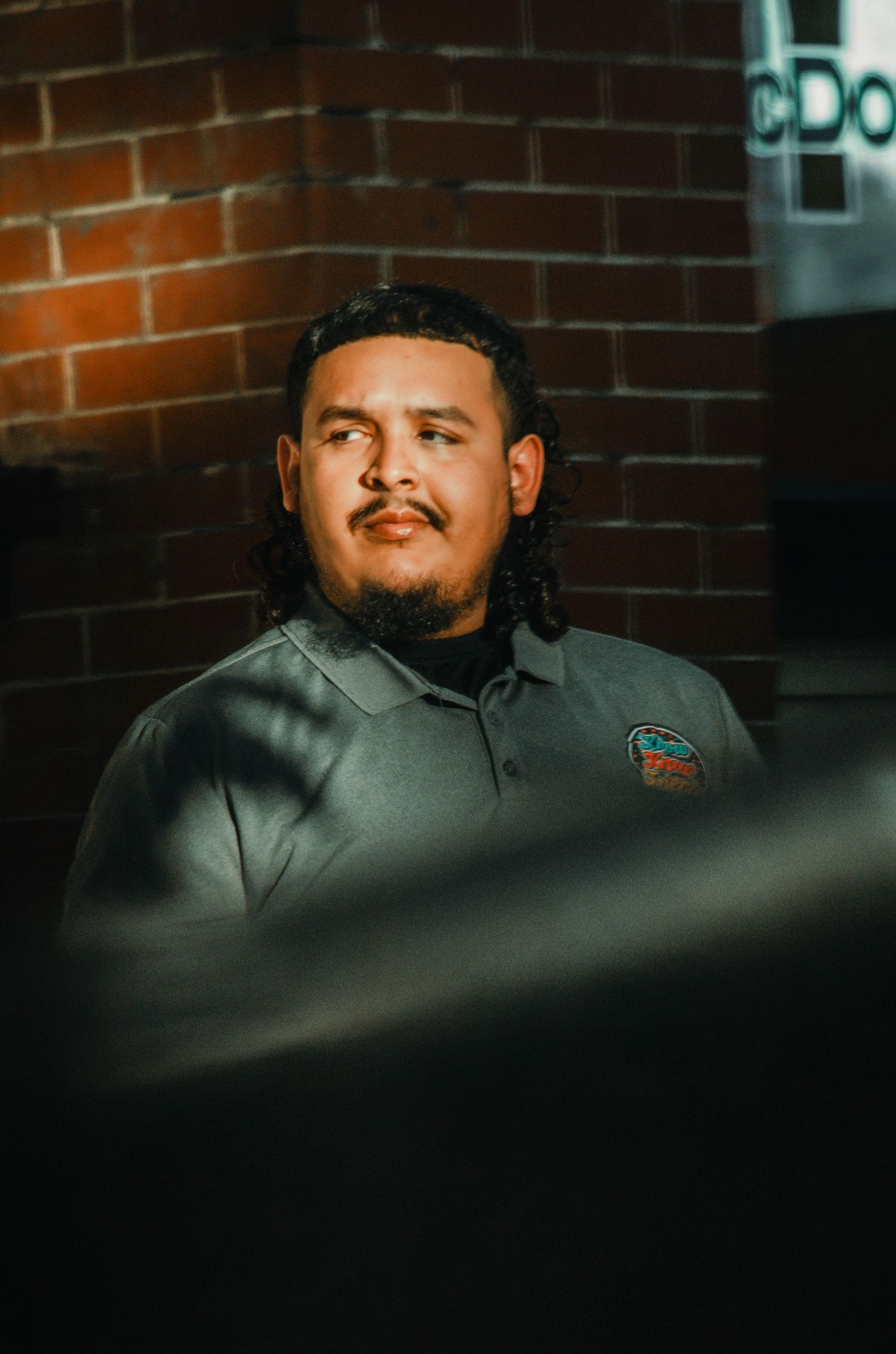 Close-up portrait of a Hispanic man with long hair and goatee wearing a gray polo shirt near a brick wall.