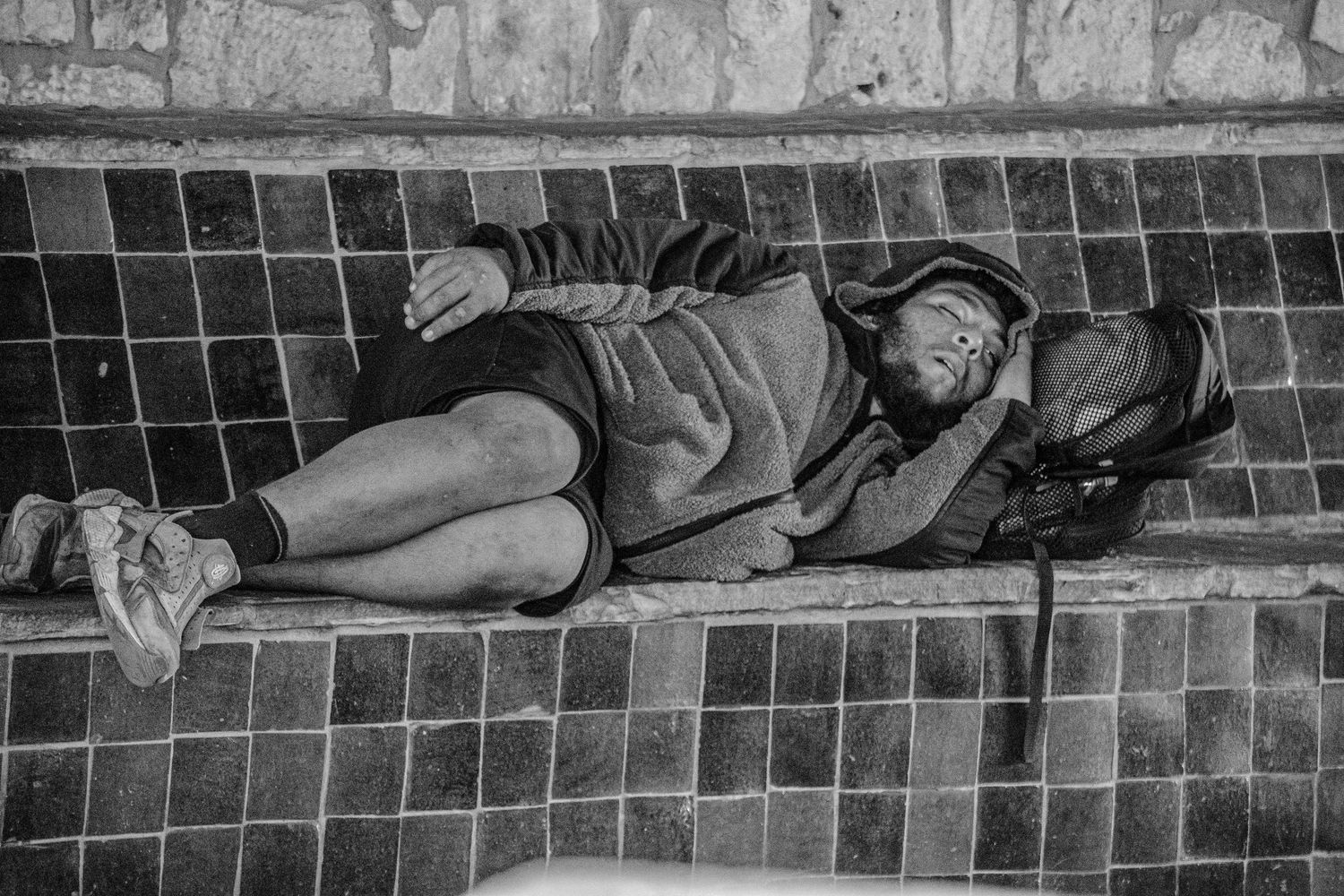 Black and white photo of a homeless person sleeping on a tiled bench outdoors.
