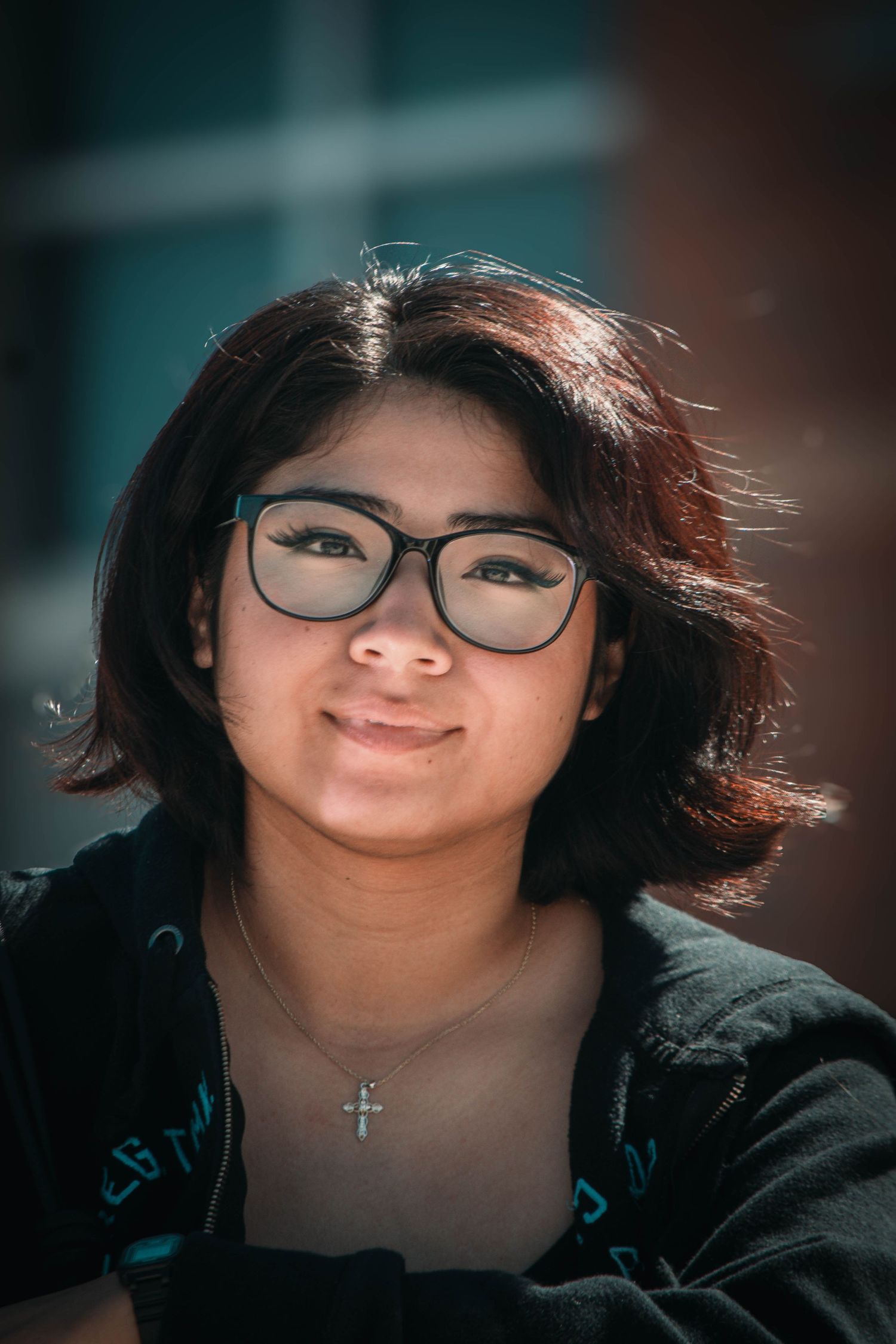 Young woman with short dark hair, glasses, and a cross necklace smiling outdoors in sunlight.