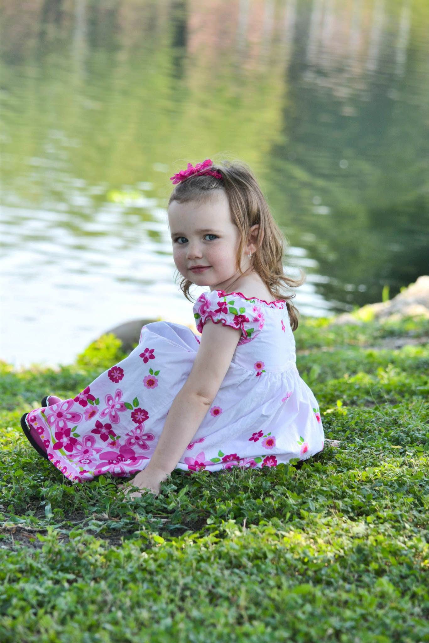 Adorable toddler girl in pink floral dress sitting on grass near a pond.