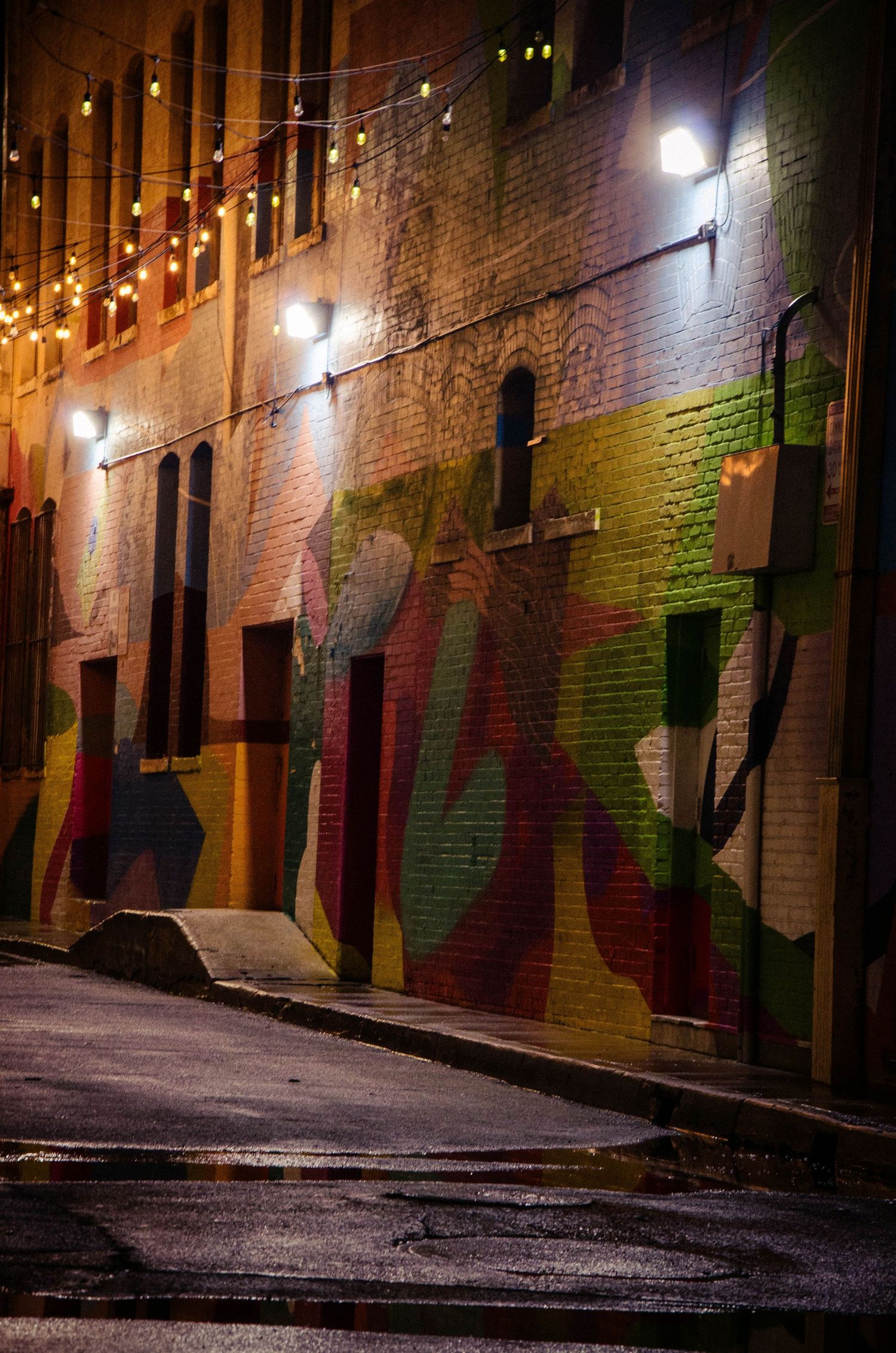 Colorful graffiti mural on brick wall in a wet urban alley illuminated by string lights at night.