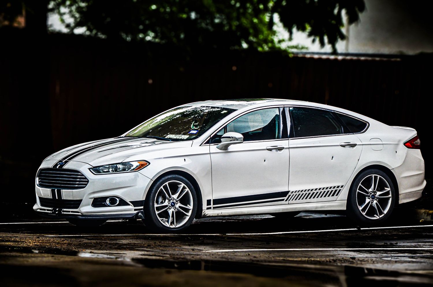A sleek white Ford Fusion sedan with black racing stripes parked on a wet surface in dramatic lighting.