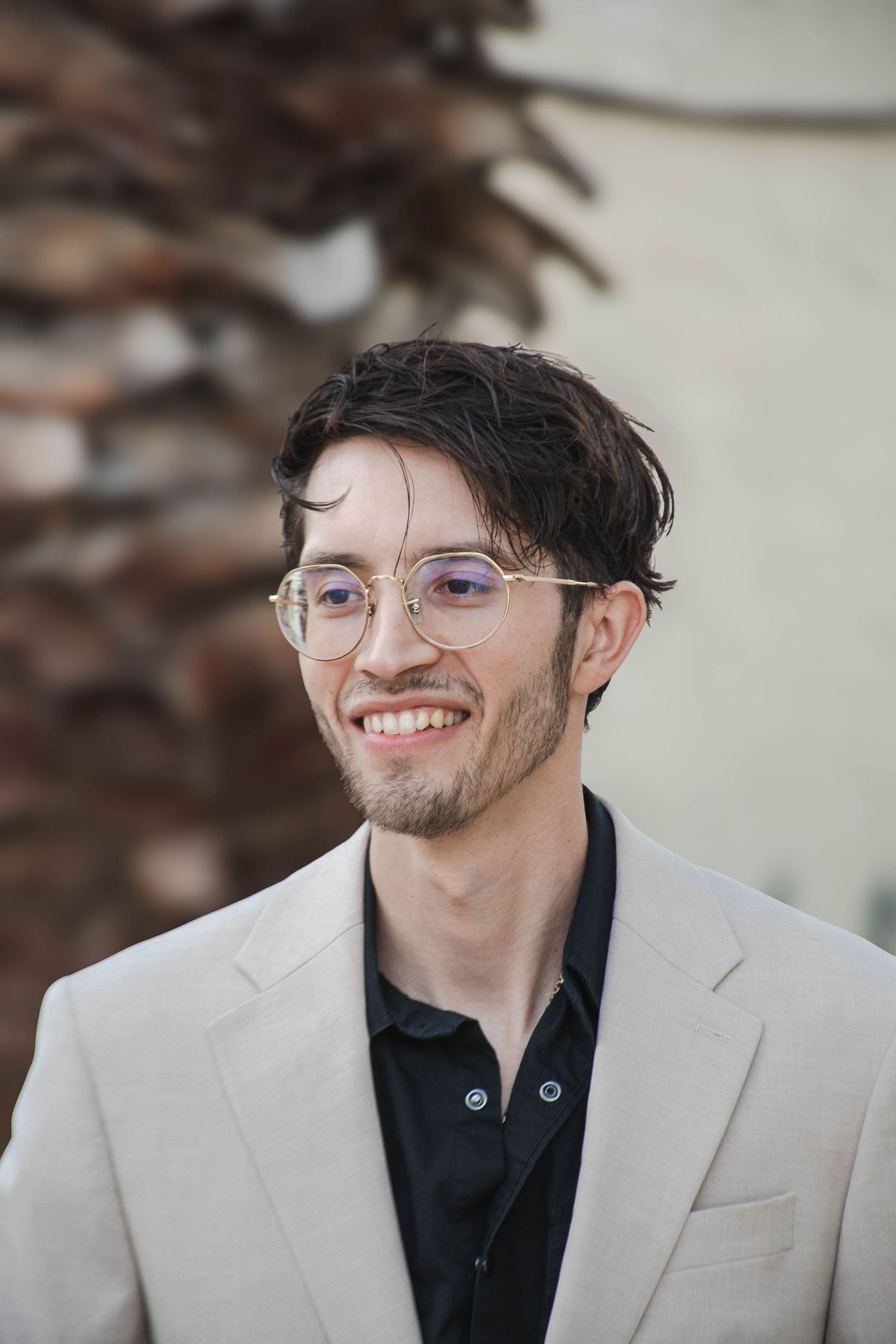 Smiling young groom with glasses and dark hair wears a beige suit and black shirt on his wedding day.