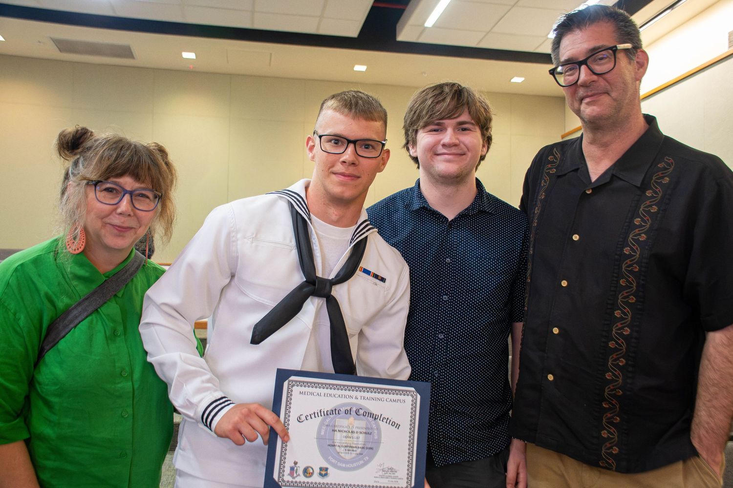 Navy sailor in dress whites holds Certificate of Completion surrounded by proud family members.