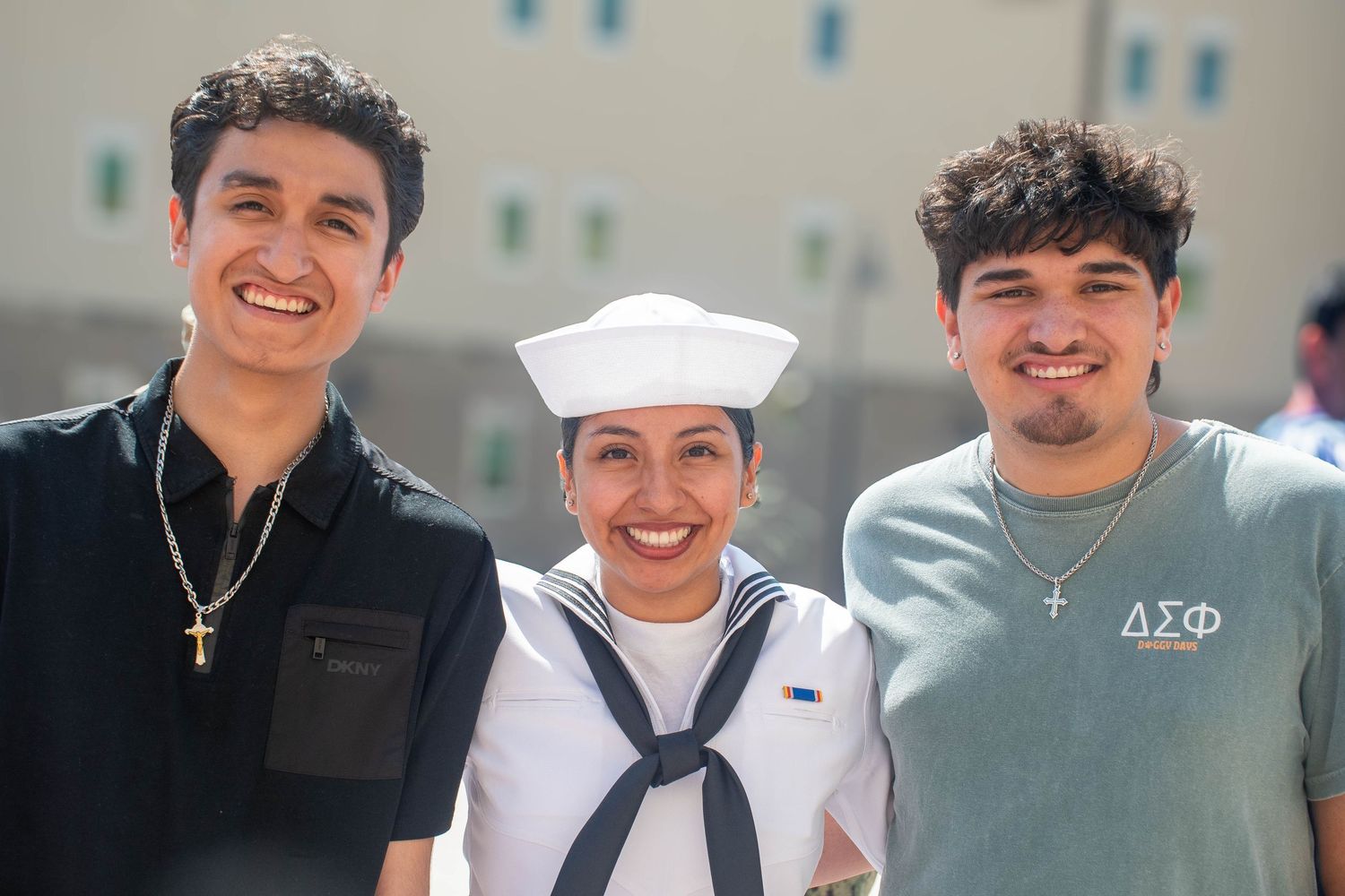 Smiling Navy sailor in dress whites stands between two young men at a military graduation event.