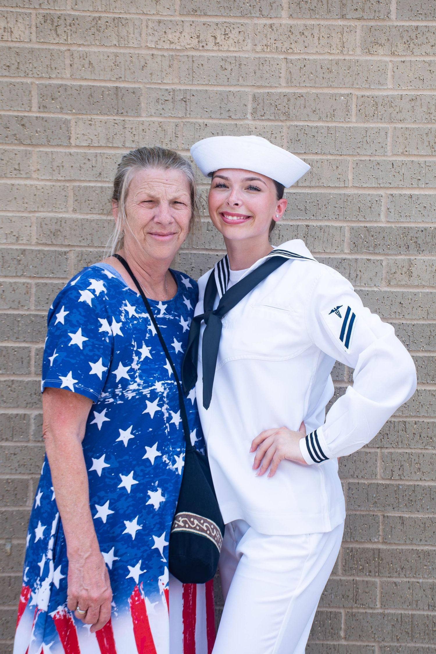 Female Navy sailor in white uniform poses with older woman in American flag dress against brick wall.