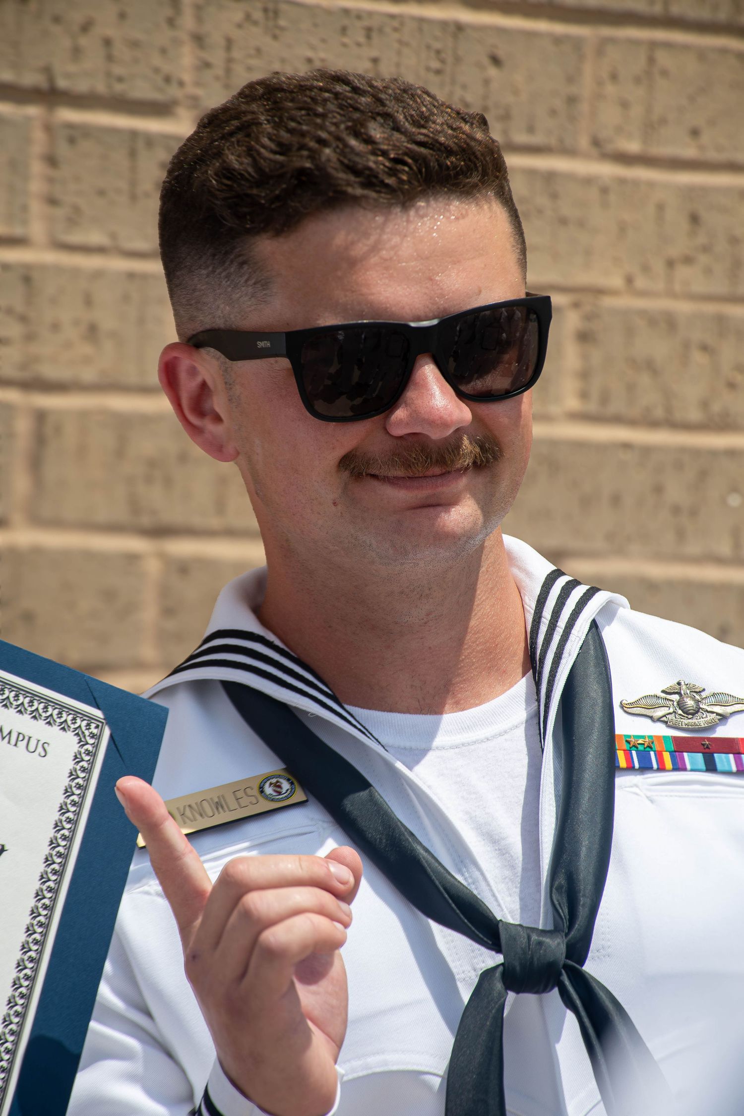 Male US Navy sailor in white uniform wearing sunglasses holding a certificate outdoors.