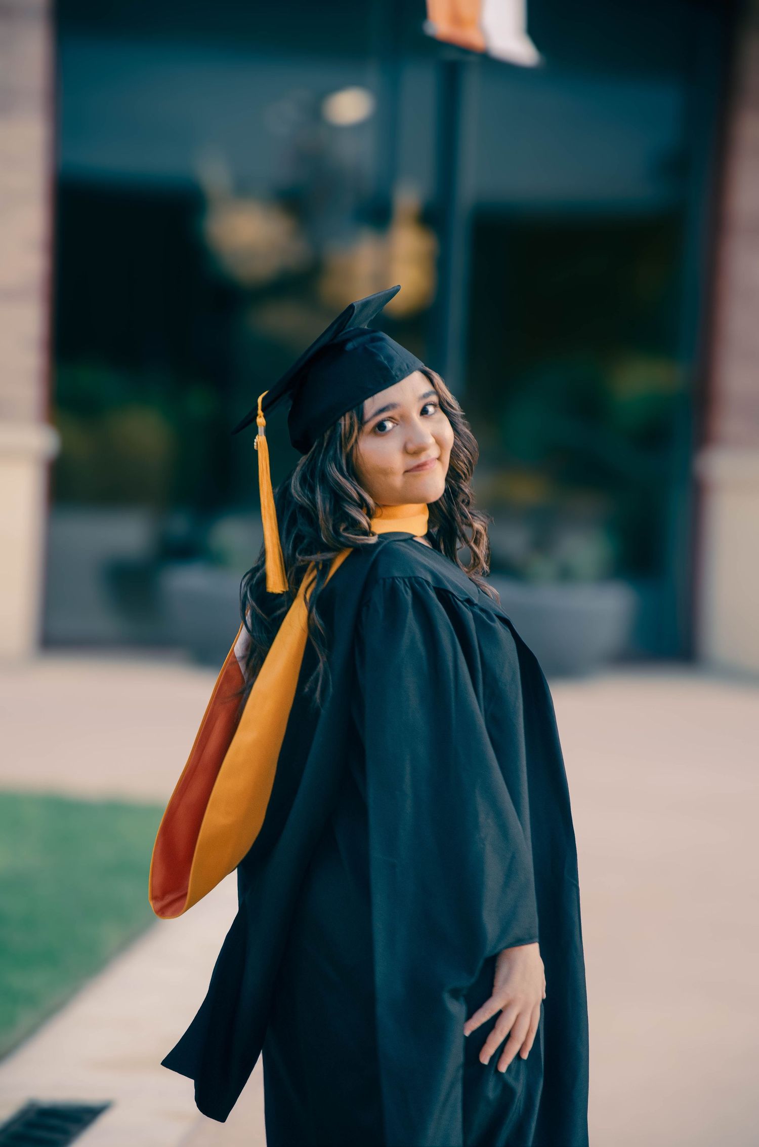 Graduate in black cap and gown with gold tassel posing outdoors at university campus.