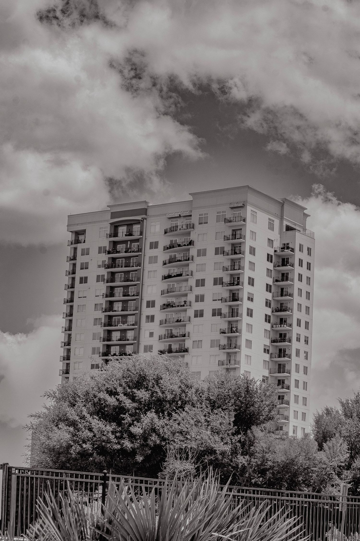 Black and white image of a tall multi-story residential apartment building against a dramatic cloudy sky.