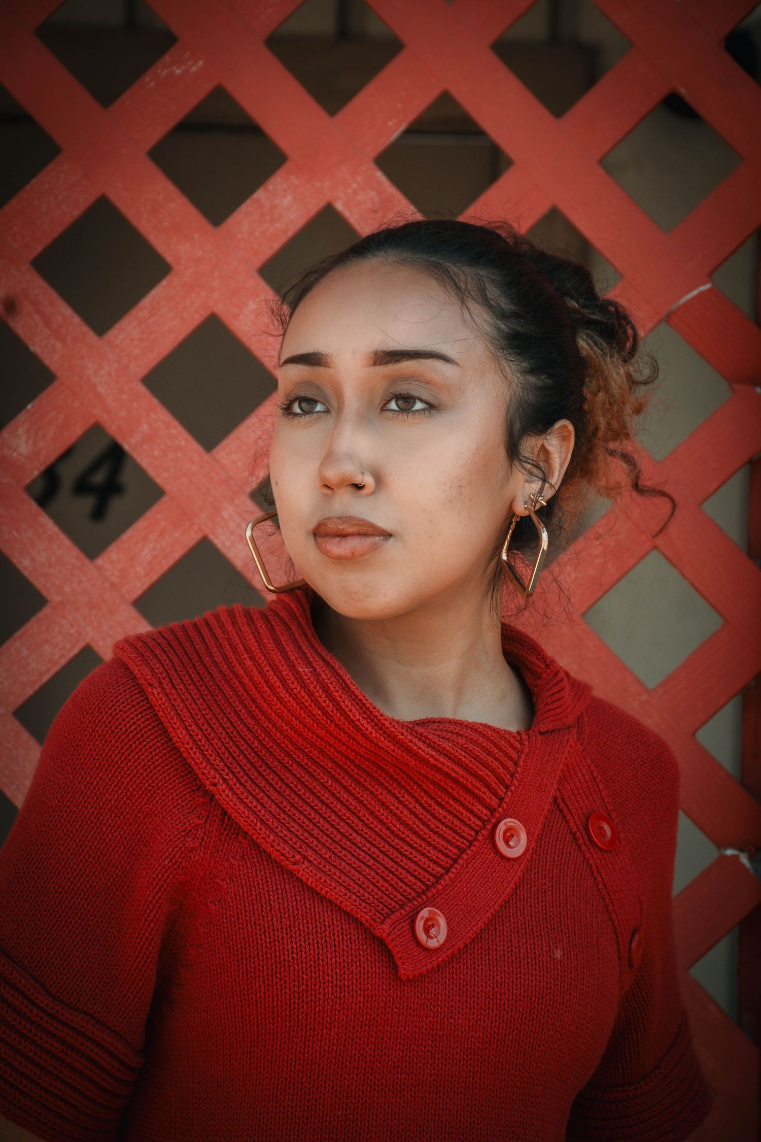 Young woman in red knit sweater with gold hoop earrings poses against a red lattice fence backdrop.