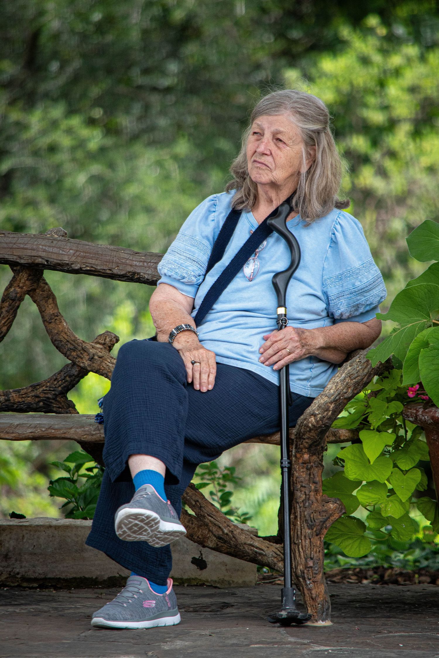 Elderly woman with gray hair sitting on rustic bench holding walking cane in park.