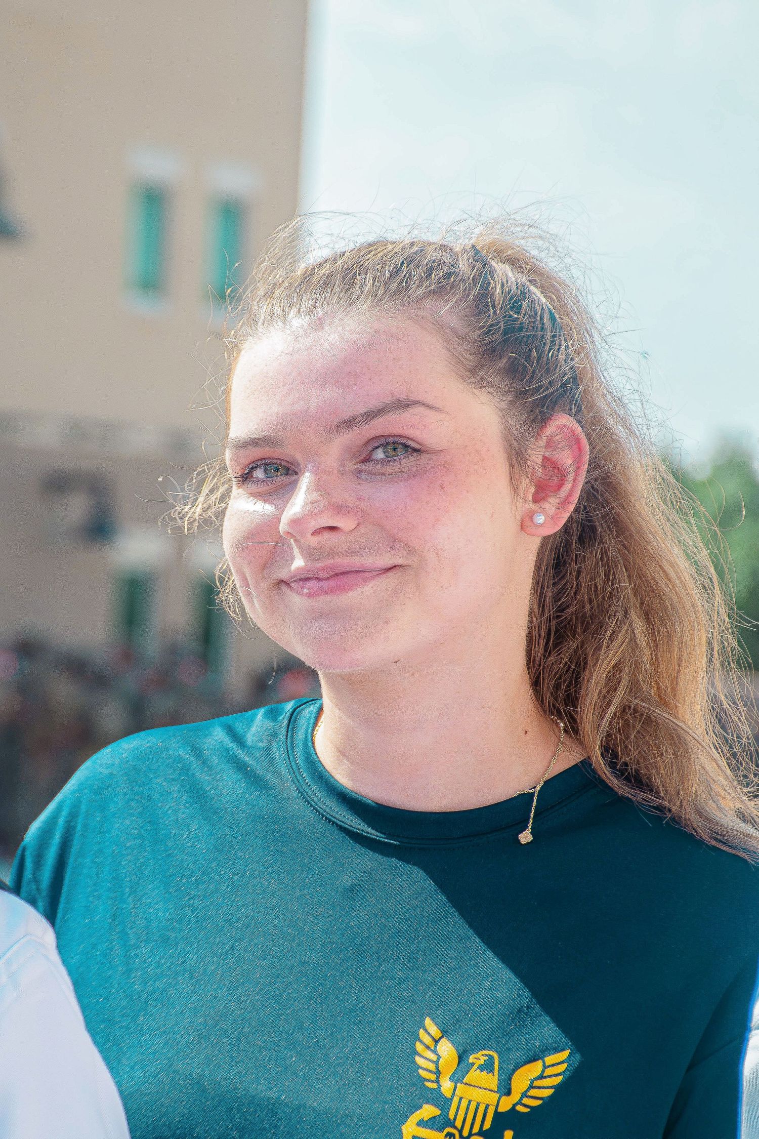 Smiling young woman with ponytail wearing a teal t-shirt with gold eagle logo outdoors.