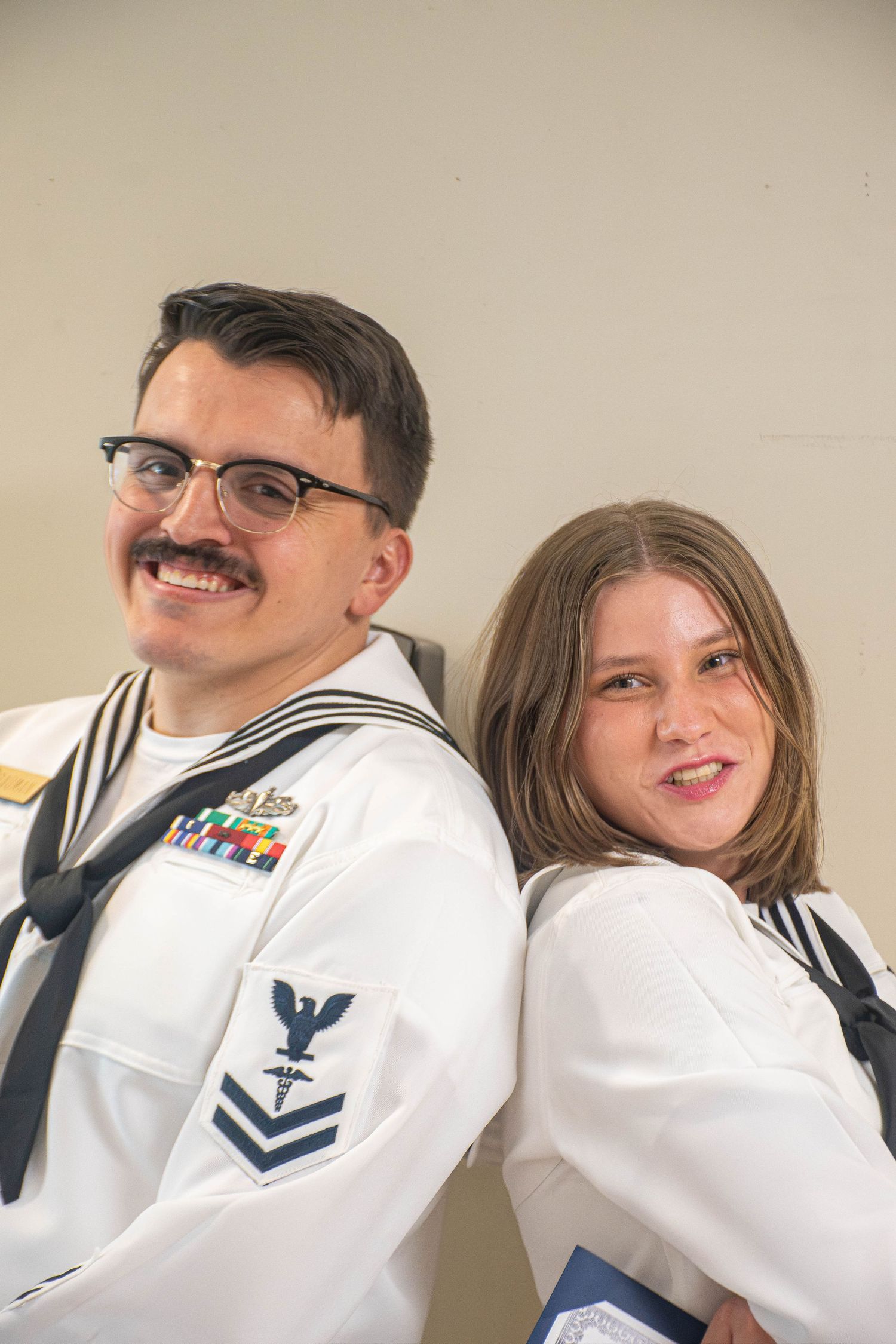 Two Navy sailors in dress white uniforms pose back-to-back smiling indoors at military event.