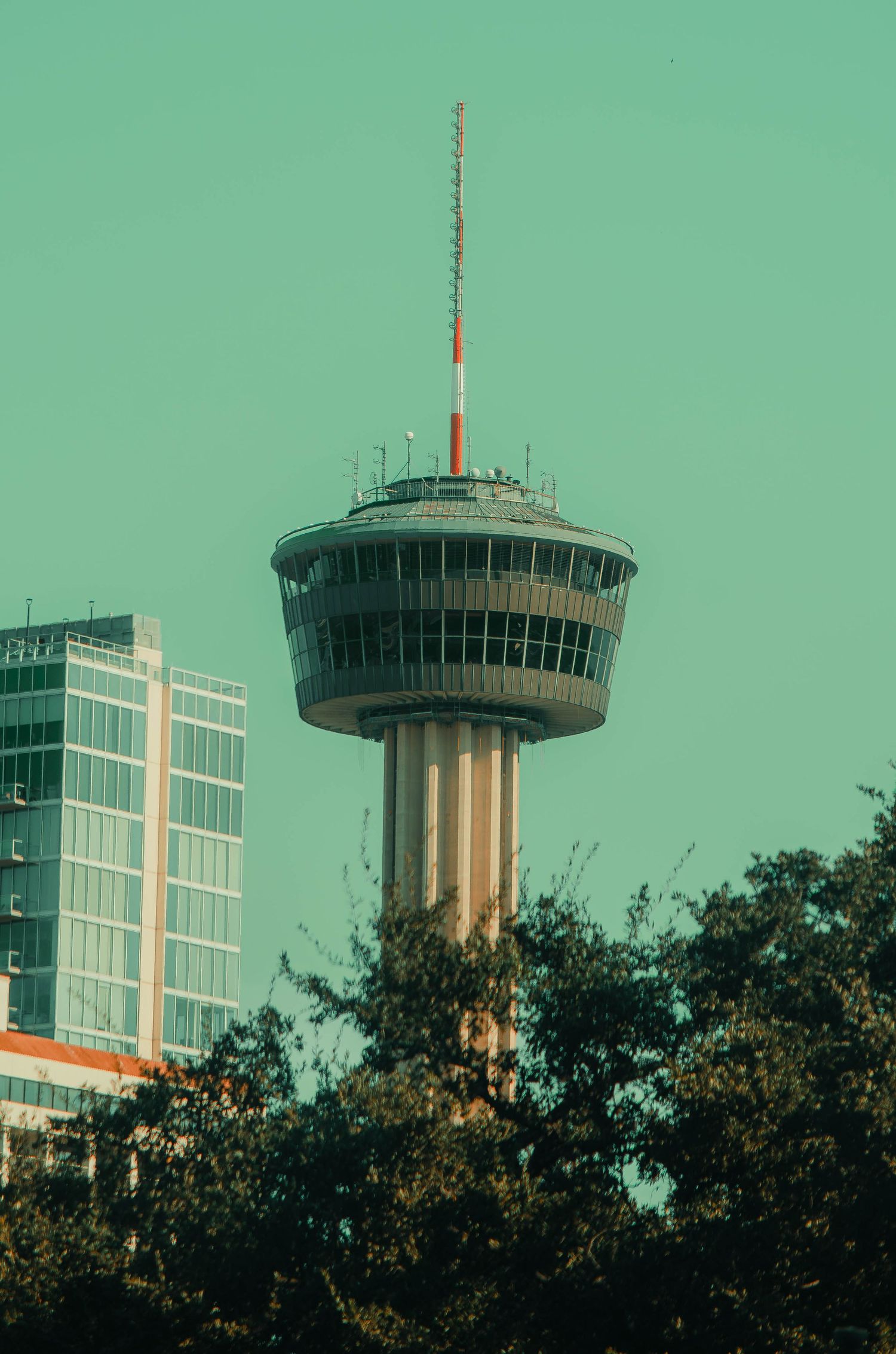 Tower of the Americas observation tower rises above green trees against a teal sky in San Antonio.