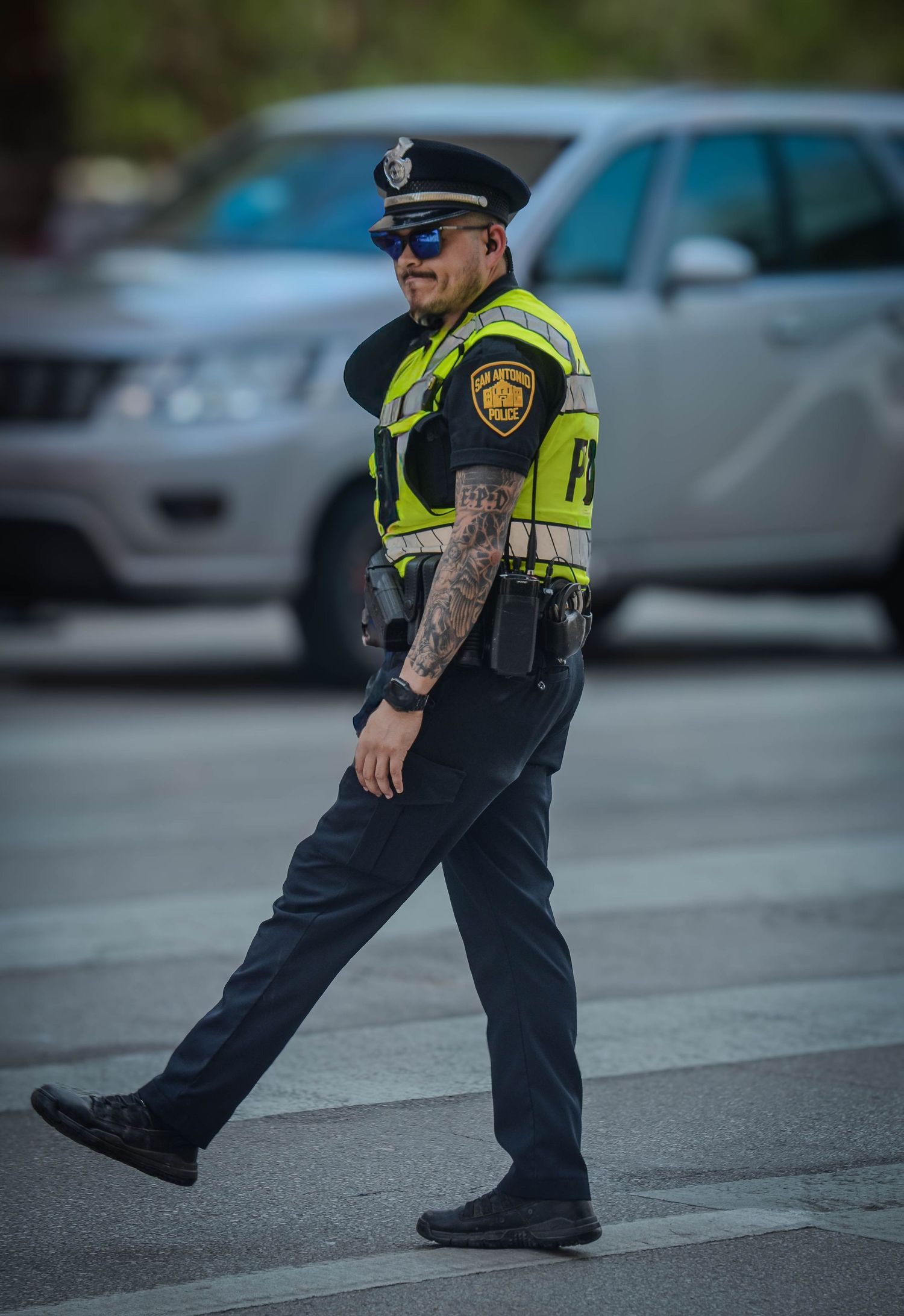 Tattooed police officer in high-visibility vest and blue uniform walking across street.