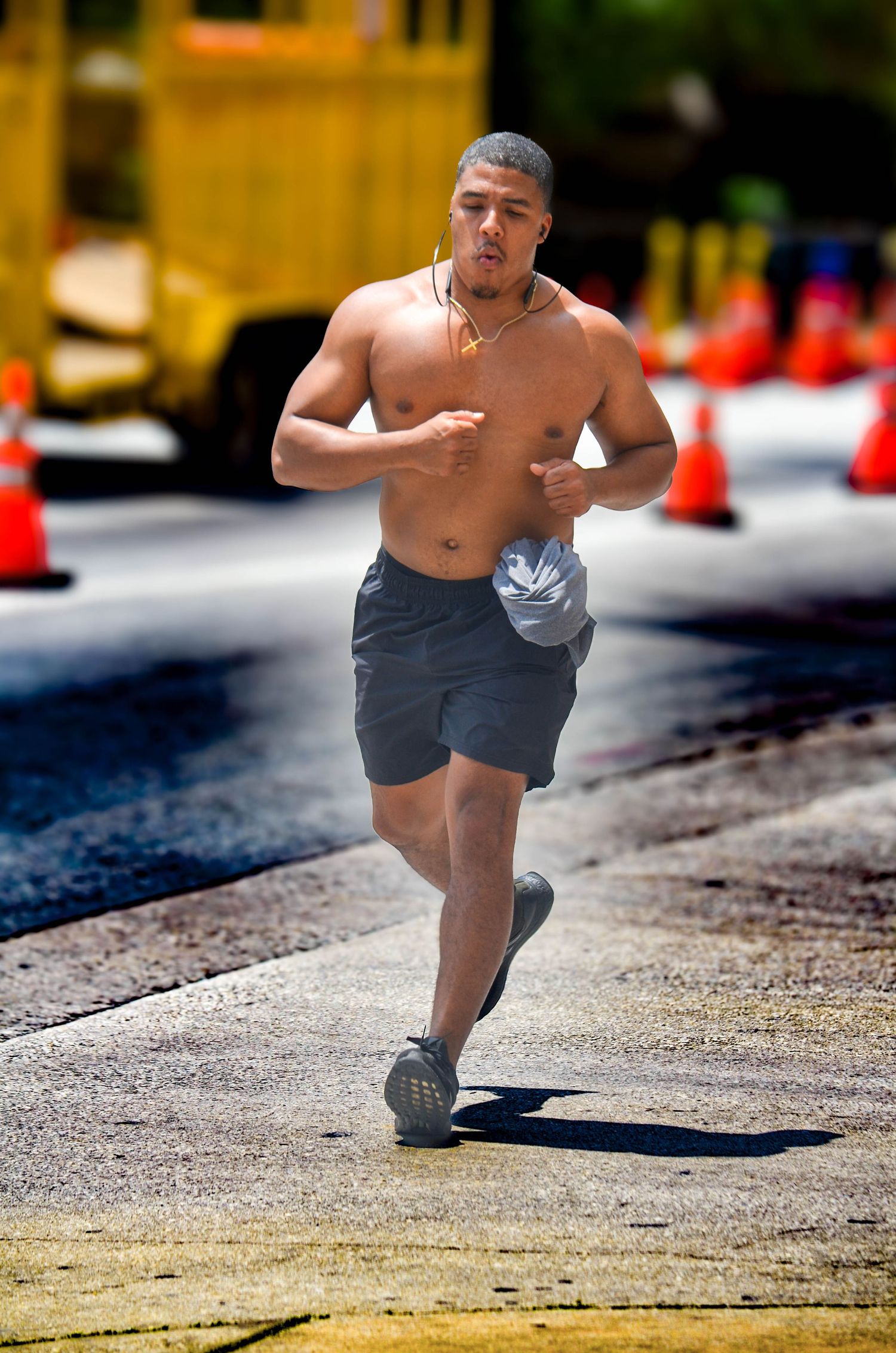 Shirtless muscular man running outdoors near orange traffic cones on a sunny day.