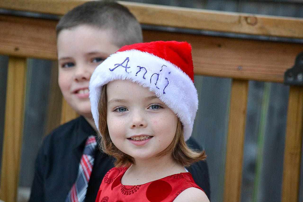 Smiling little girl wearing a personalized Santa hat with a boy behind her.