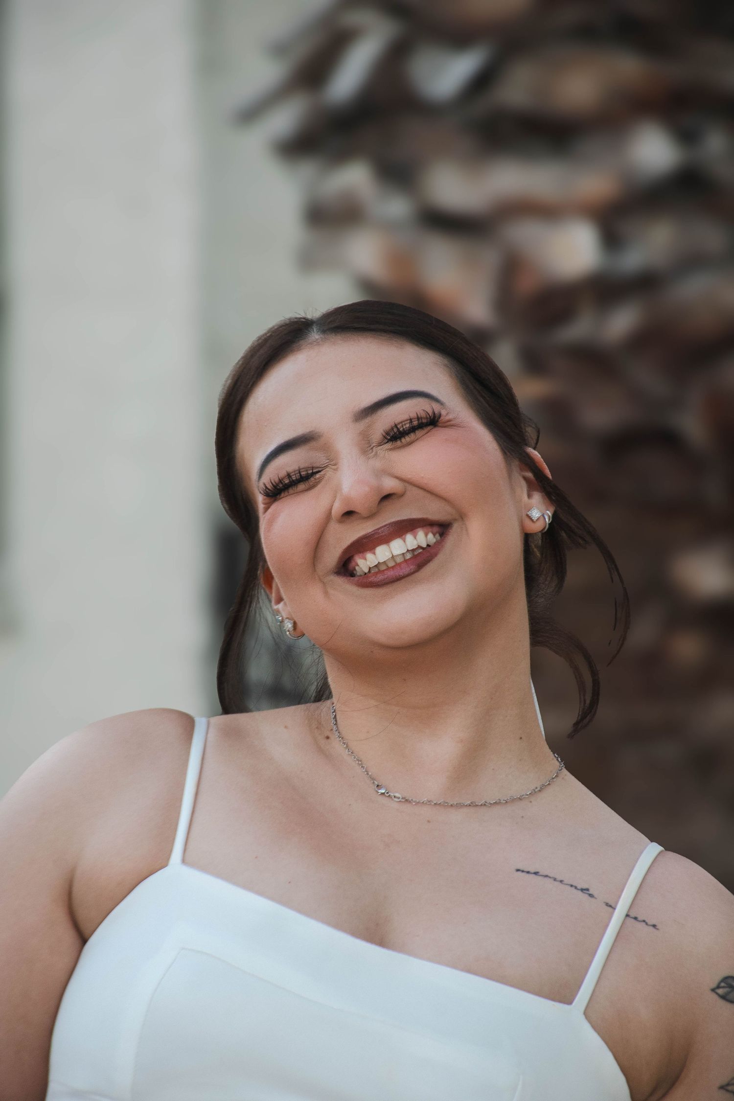 Radiant bride in white dress laughs joyfully with eyes closed near a palm tree on her wedding day.
