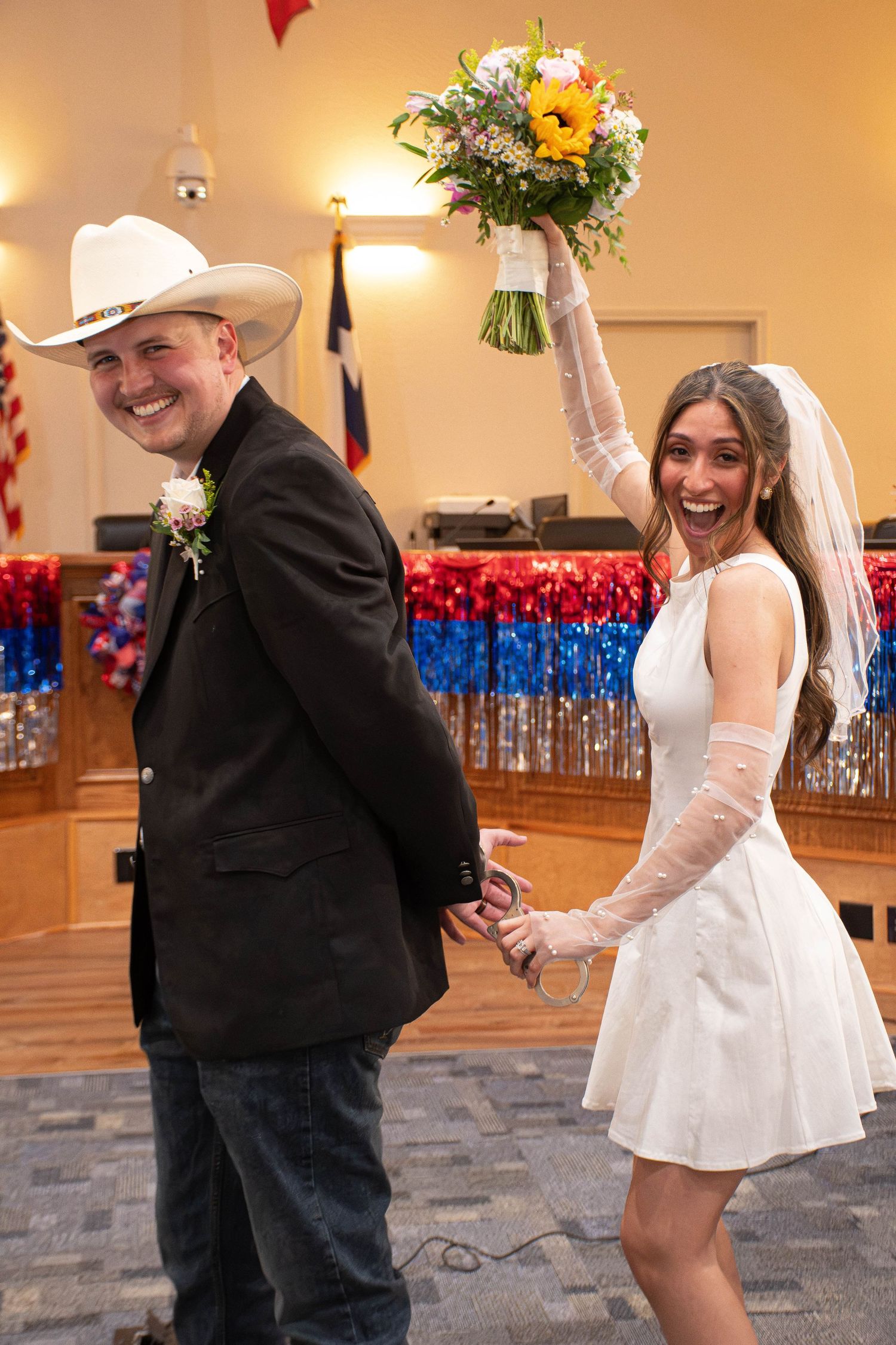 Joyful bride in short white dress holds bouquet aloft while groom in cowboy hat and black suit smiles at wedding reception.