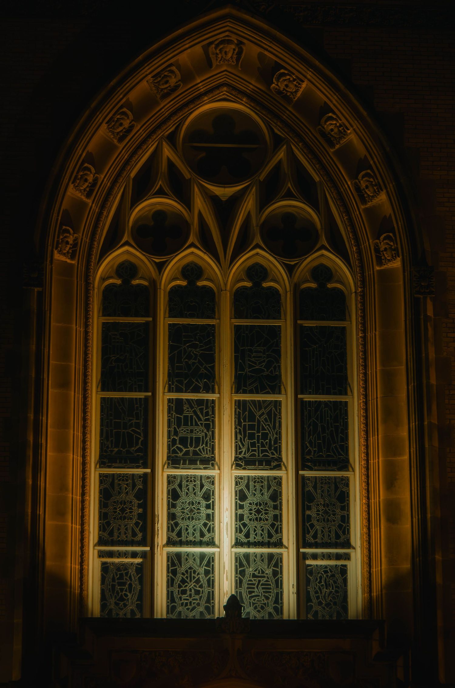 Gothic cathedral window with intricate tracery illuminated by warm golden light at night.