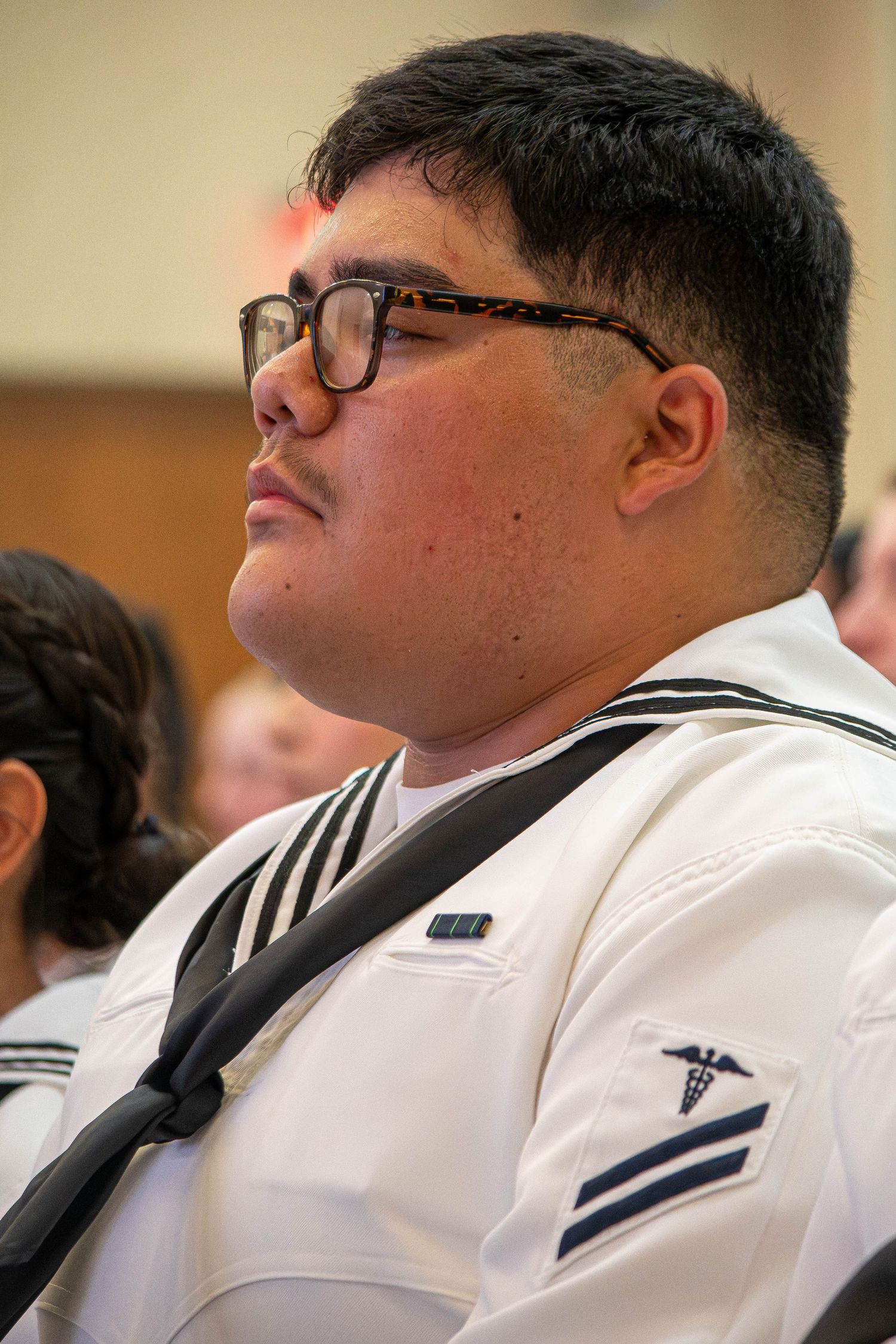 Male Navy sailor wearing glasses in white uniform with medical corps insignia looking upward.